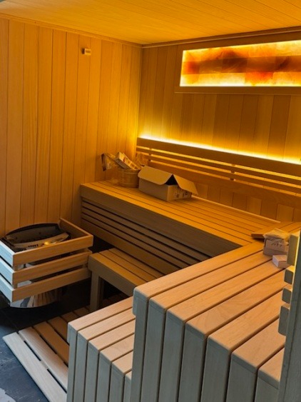 Jasne drewno w saunie z oświetleniem LED i panelem solnym. Widoczne ławy, piec i akcesoria do sauny. Przytulne, relaksujące wnętrze.