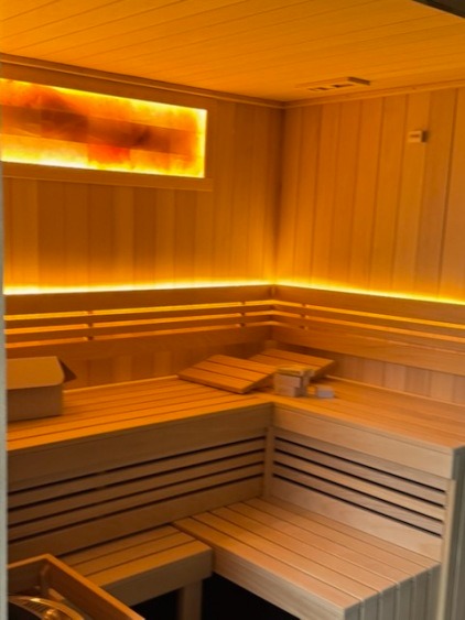 Jasne drewno i ciepłe oświetlenie tworzą przytulne wnętrze sauny z ławkami w układzie litery L i podświetlanym panelem solnym na ścianie.