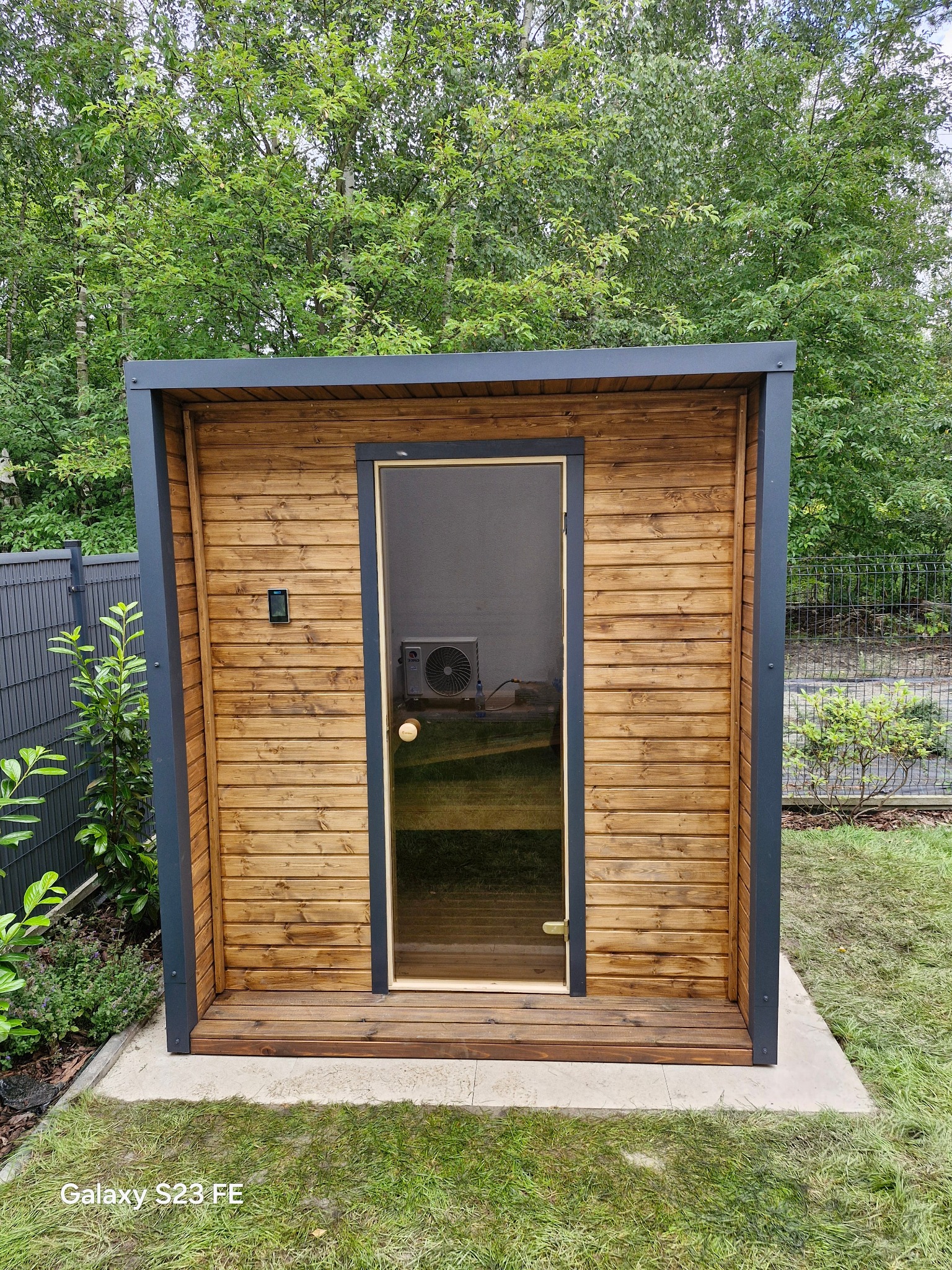 Drewniana sauna ogrodowa z przeszklonymi drzwiami na betonowej podstawie, otoczona zielenią, z widocznym klimatyzatorem wewnątrz.