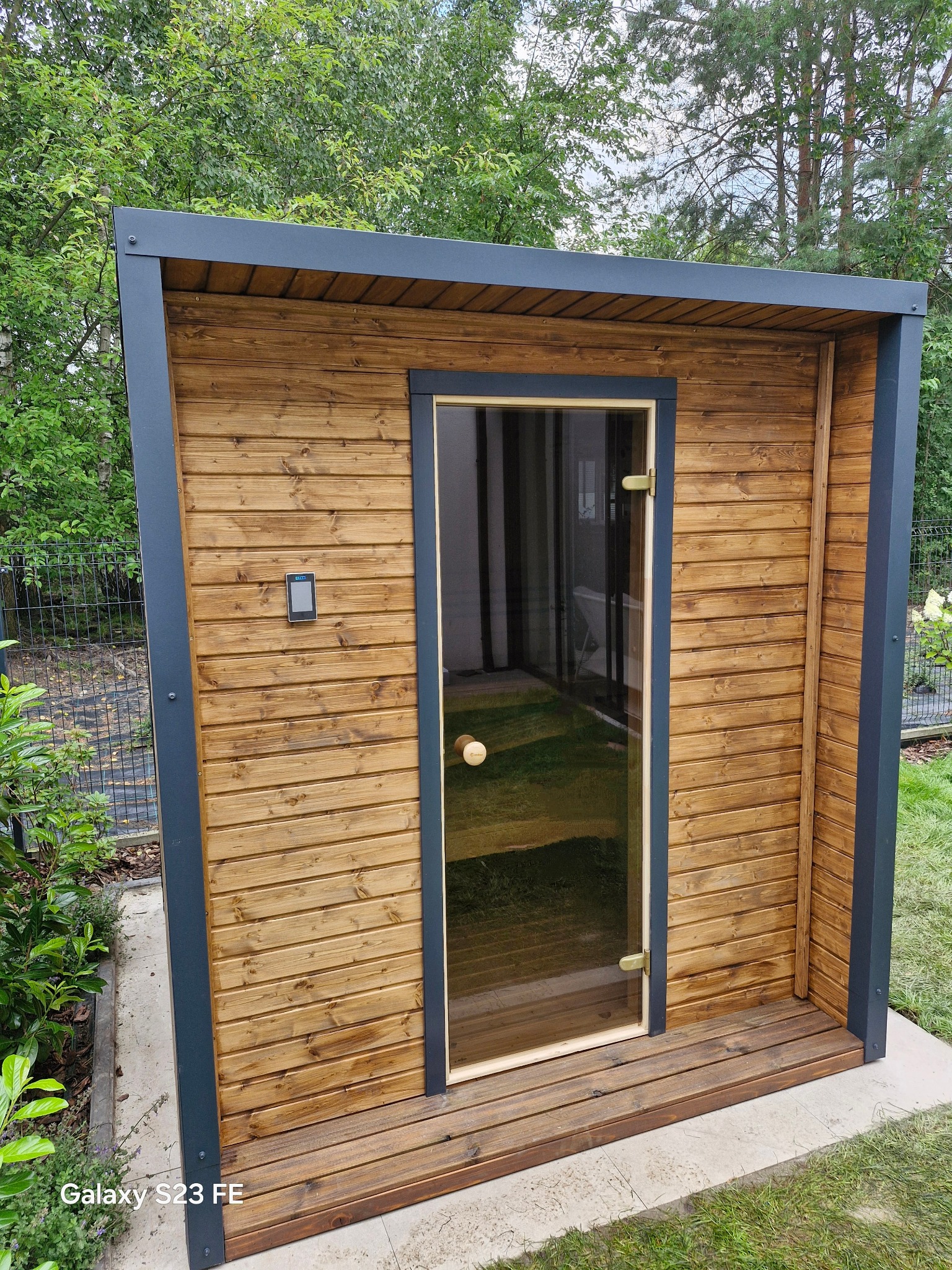 Drewniana sauna ogrodowa z przeszklonymi drzwiami, otoczona zielenią. Widoczne deski z wyraźnym usłojeniem i metalowe elementy konstrukcyjne w kolorze grafitowym.