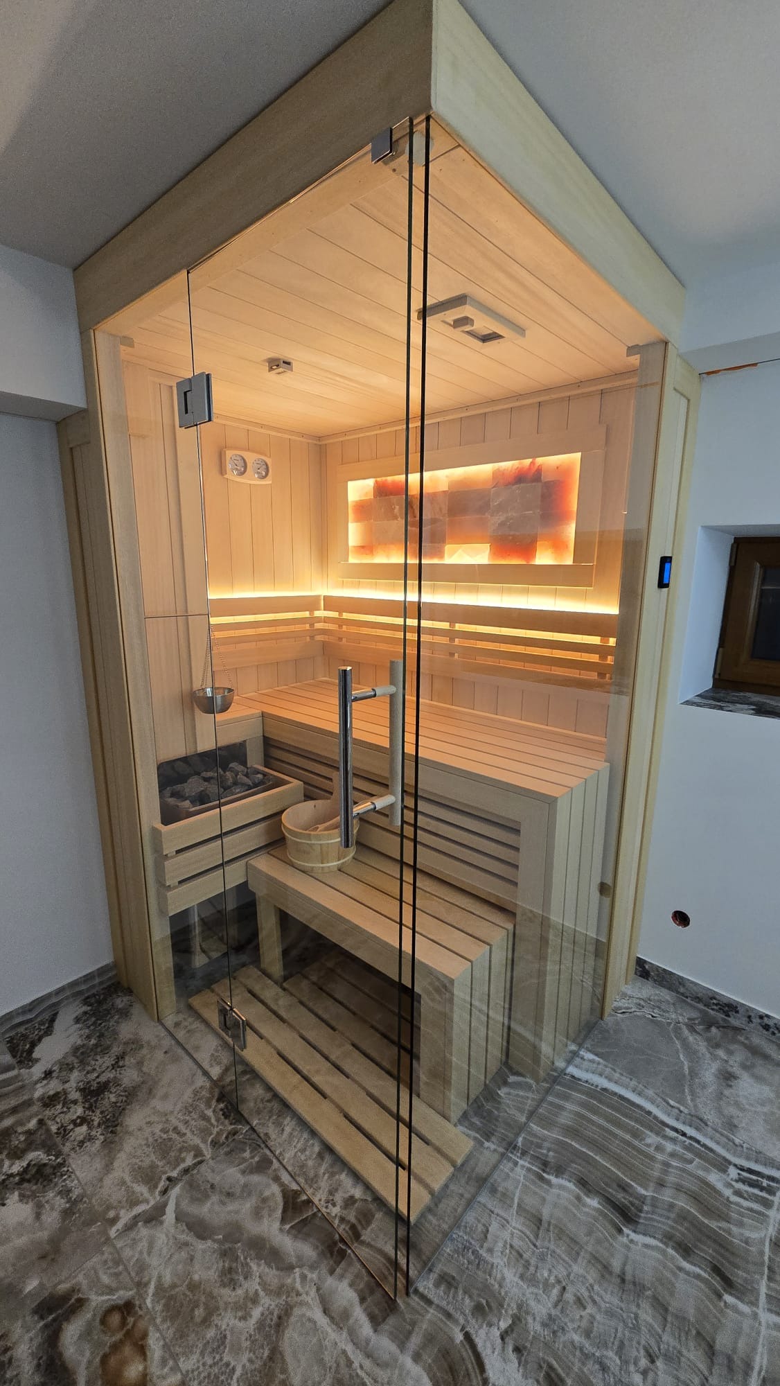 Nowoczesna sauna z przeszklonymi ścianami, drewnianymi ławkami i panelem solnym podświetlonym LED, widok z zewnątrz na marmurową podłogę.