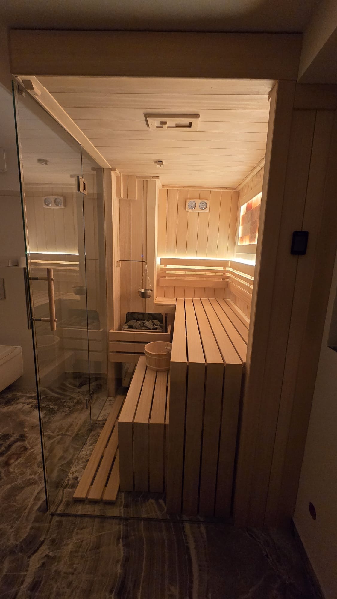 Nowoczesna sauna z przeszklonym wejściem, drewnianymi ławkami i kamienną ścianą solną. Subtelne oświetlenie LED podkreśla ciepło drewna i tworzy relaksującą atmosferę.