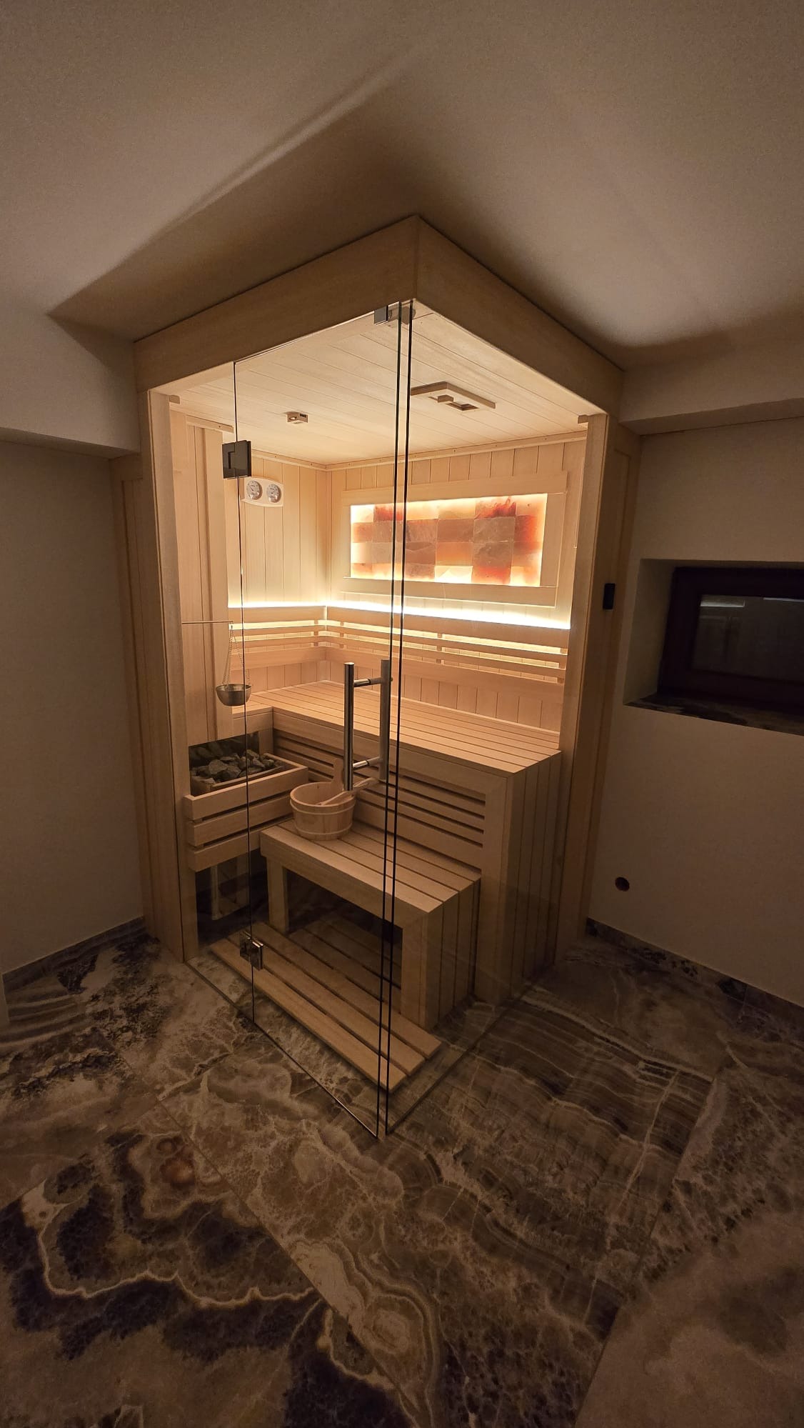 Nowoczesna sauna z przeszklonymi drzwiami, drewnianym wykończeniem i podświetlanymi panelami solnymi. Widoczny kamienny podest i akcesoria do sauny.
