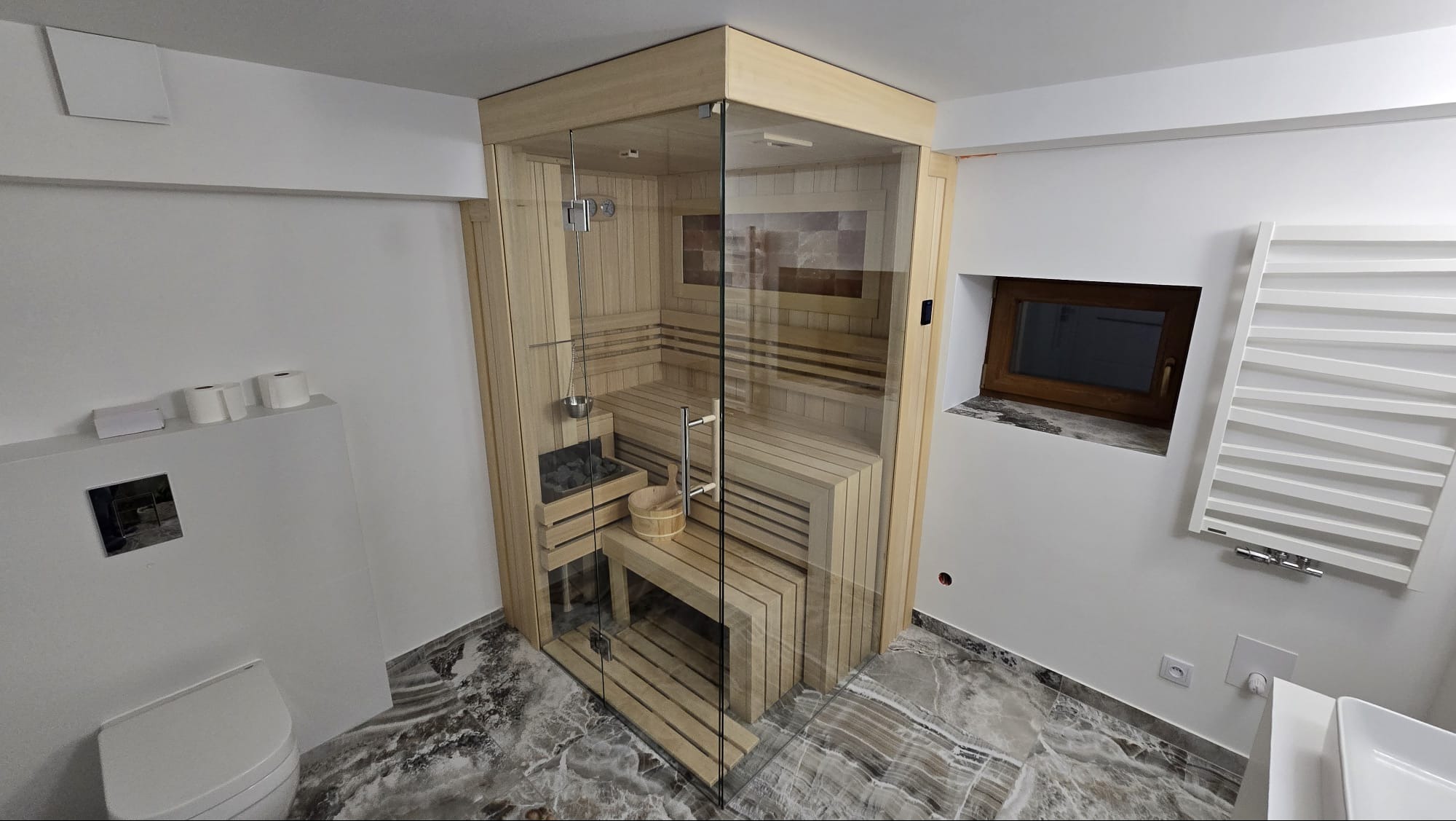 Nowoczesna, przeszklona kabina sauny z jasnego drewna w łazience z marmurową podłogą. Widoczne detale: piec z kamieniami, drewniane ławki, wiadro i czerpak.