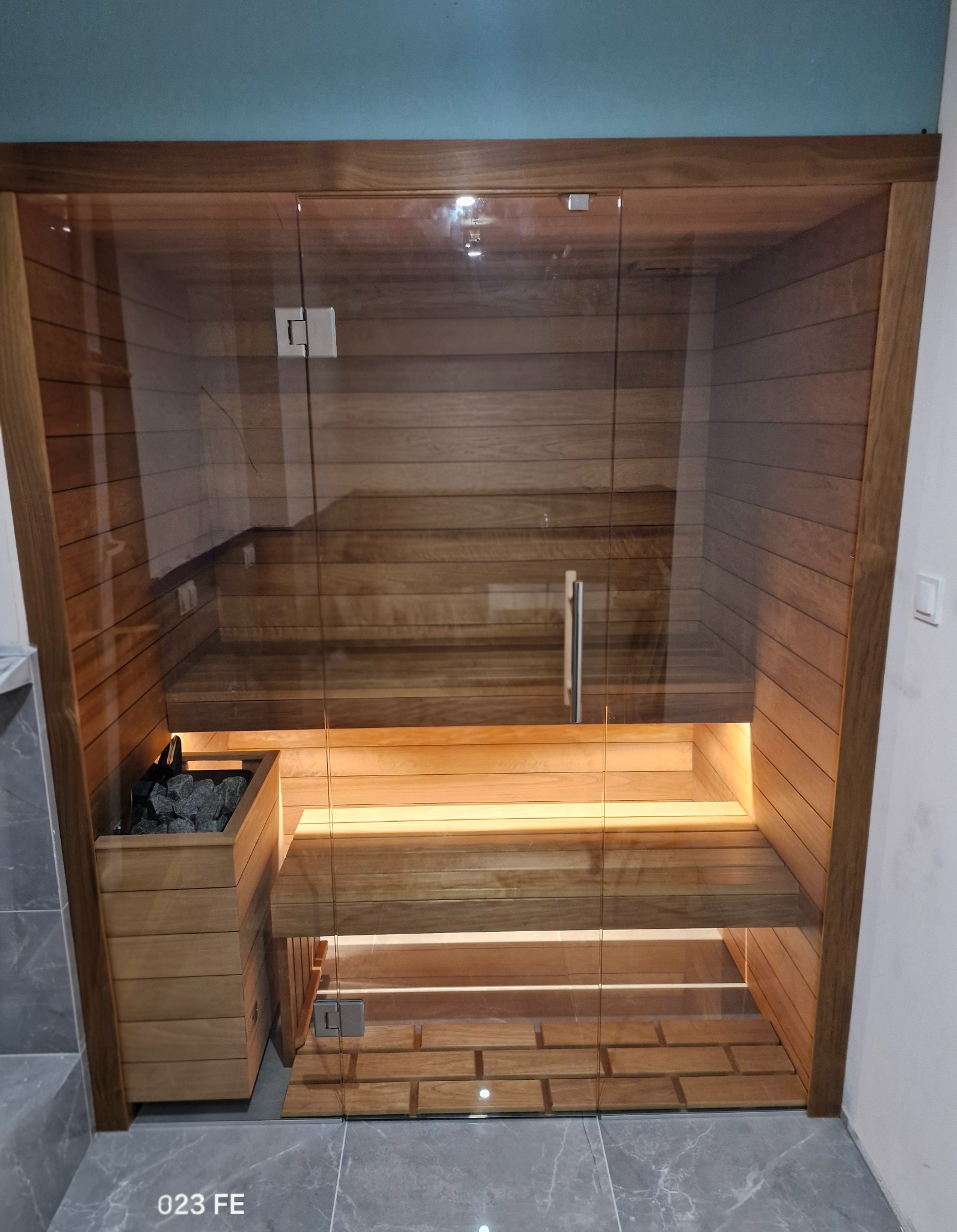 Nowoczesna, przeszklona kabina sauny z drewnianym wykończeniem i oświetleniem LED, widok z przodu, z detalami kamieni w pojemniku i szklanych drzwi.