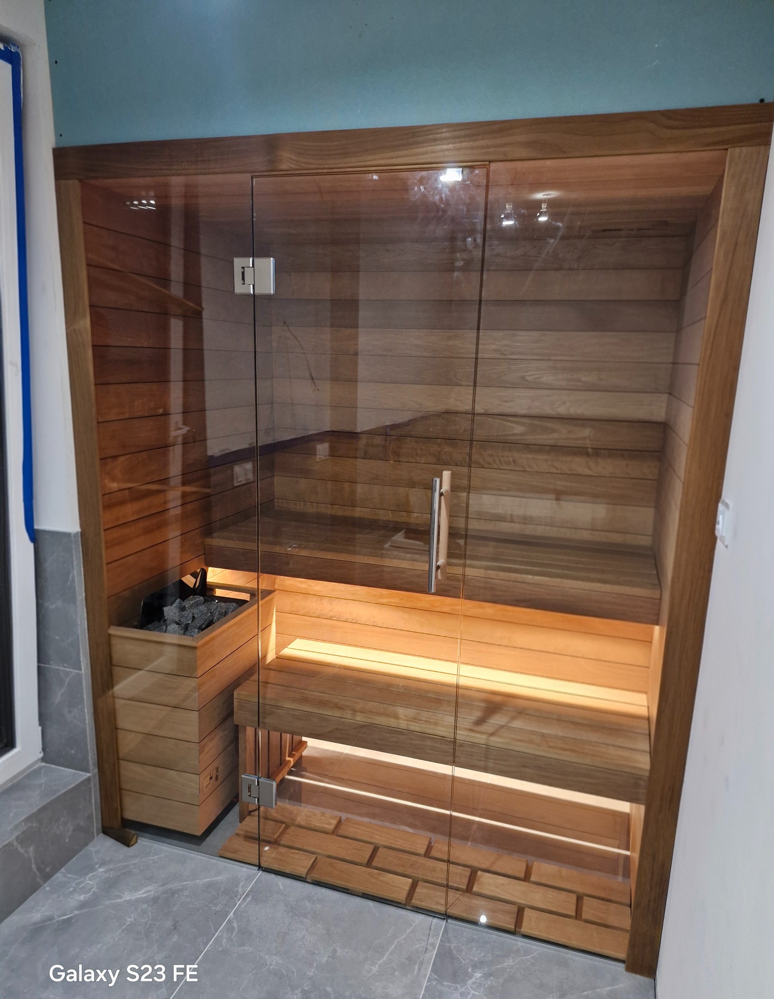 Nowoczesna sauna z przeszklonymi drzwiami i drewnianym wykończeniem. Widoczne kamienie w piecu i podświetlone ławki. Minimalistyczny design, jasne drewno.