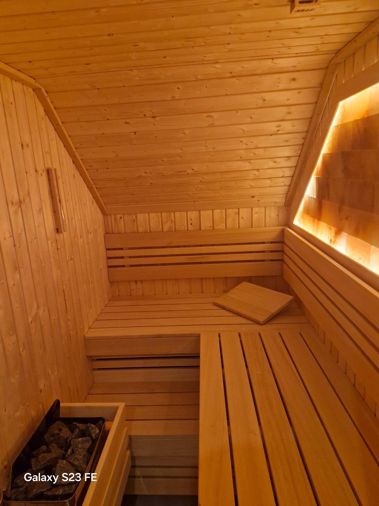 Wnętrze drewnianej sauny z oświetleniem solnym, widok z dołu na ławy, piec z kamieniami i drewniane wykończenie ścian i sufitu. Ciepłe, relaksujące światło.