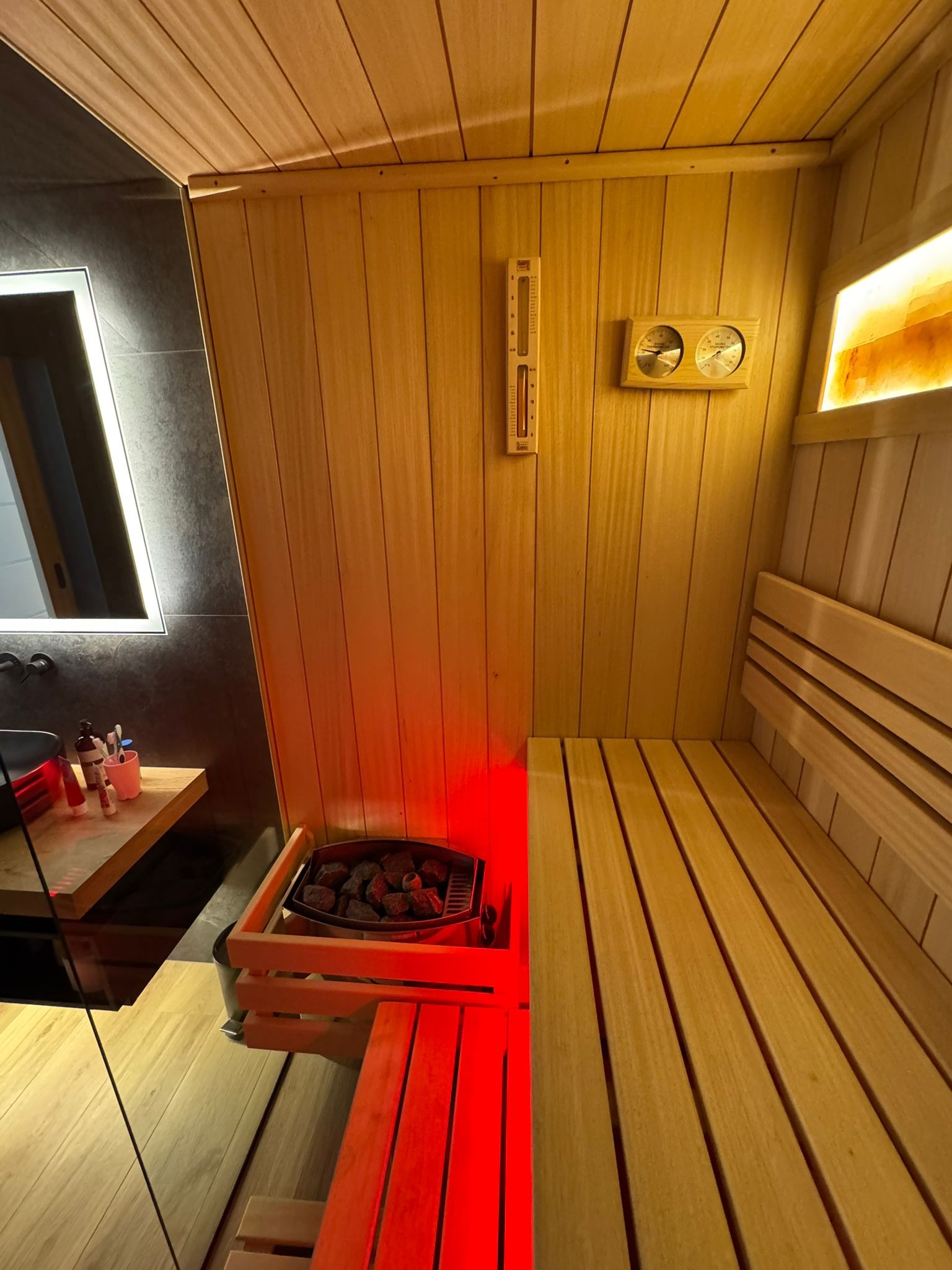Wnętrze sauny z drewnianymi ławkami, piecem z kamieniami i oświetleniem. Widoczny termometr i higrometr na ścianie. Obok sauny umywalka z lustrem.