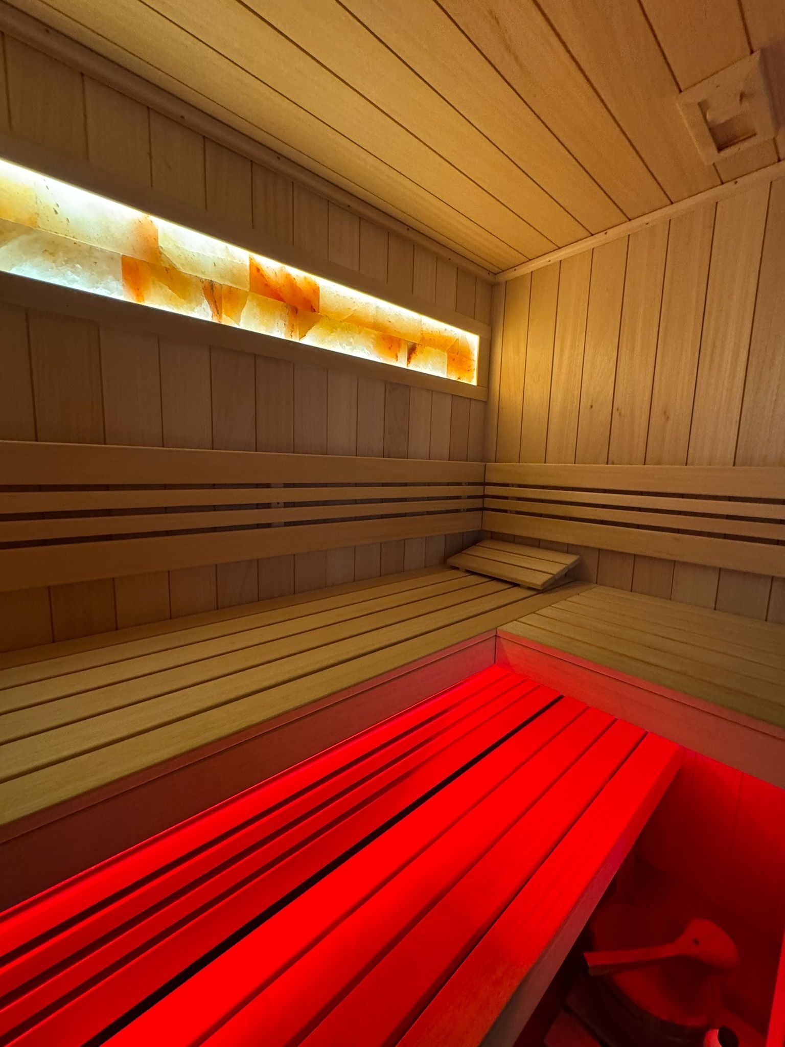 Wnętrze drewnianej sauny z podświetlanym panelem solnym i czerwonym oświetleniem pod ławkami. Widoczne drewniane ławki i oparcia, oraz akcesoria do sauny.