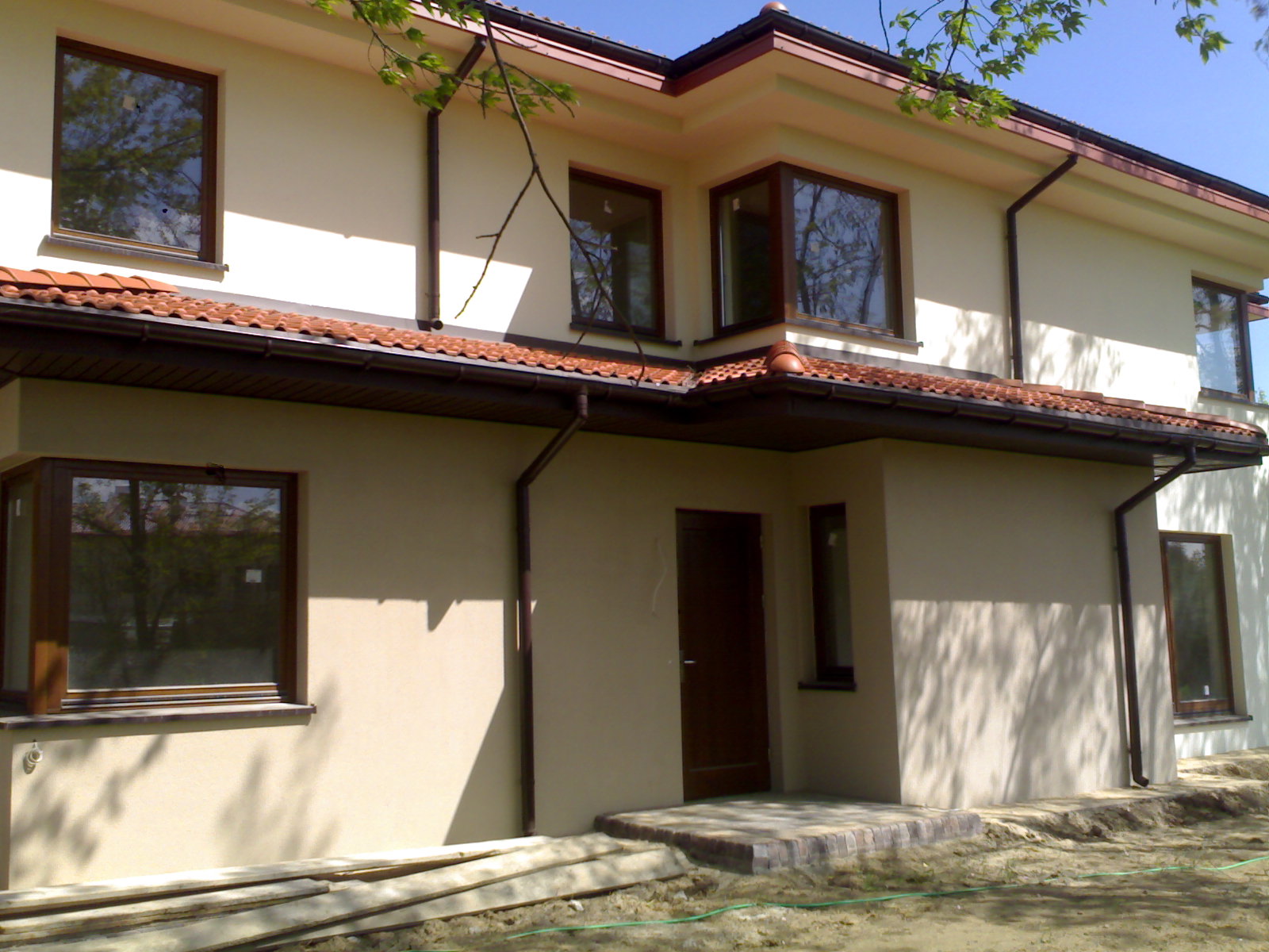 Dwukondygnacyjny dom jednorodzinny z beżową elewacją i brązową stolarką okienną, widoczny z boku w słoneczny dzień, z fragmentem dachu pokrytego czerwoną dachówką i widocznymi rynnami,...