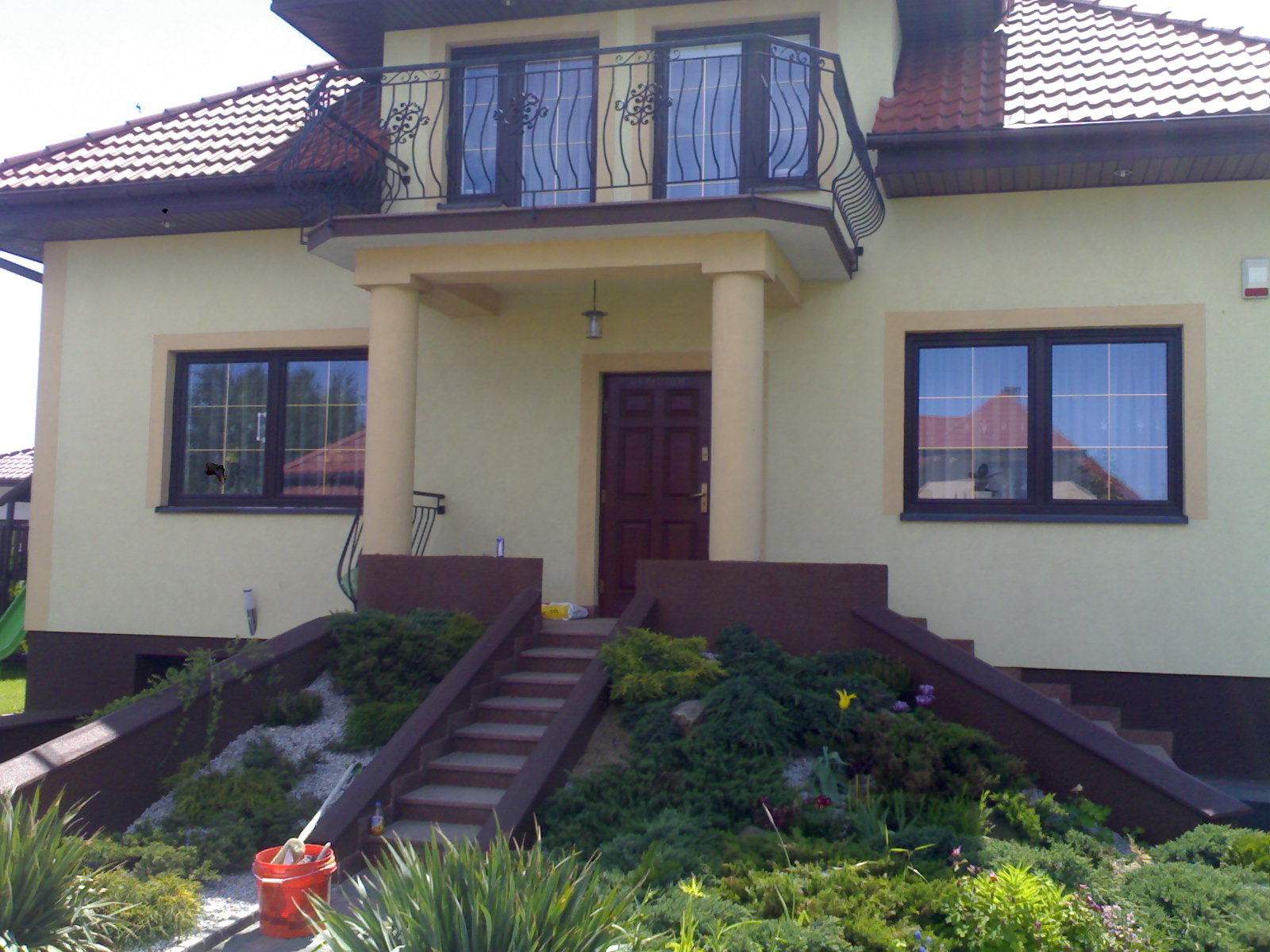 Jasna elewacja domu jednorodzinnego z brązowymi oknami i schodami wejściowymi, widoczny balkon z ozdobną balustradą i częściowo zagospodarowany ogródek przed domem.