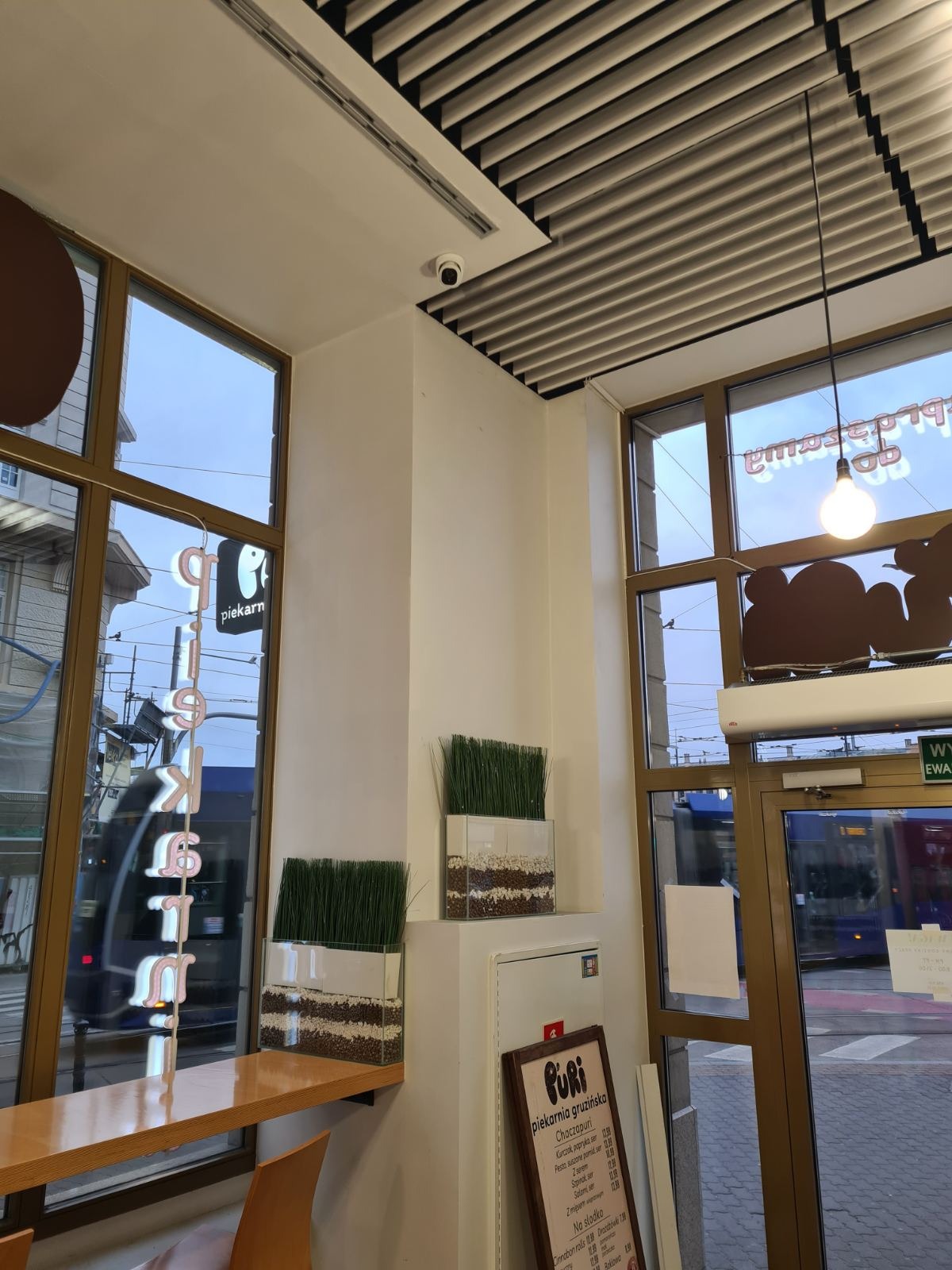 Wnętrze lokalu gastronomicznego z widoczną kamerą monitoringu na suficie, dekoracjami roślinnymi w szklanych pojemnikach, menu w ramce oraz dużymi oknami z widokiem na ulicę i przejeżdżający tramwaj.