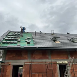 Montaż szarej dachówki na nowym budynku w trakcie budowy, widoczne rusztowania i okna dachowe, pochmurne niebo w tle.