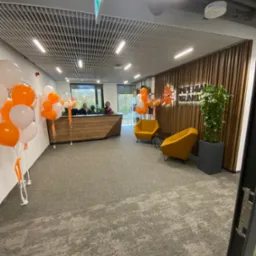 Nowoczesne biuro z recepcją, drewnianą ścianą z logo firmy Imperial Brands, dwoma żółtymi fotelami i dekoracją z białych i pomarańczowych balonów.