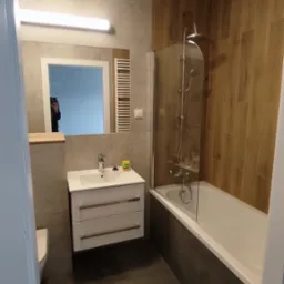 Nowoczesna łazienka po renowacji z drewnopodobnymi panelami na ścianie przy wannie, szklanym parawanem nawannowym, umywalką z szafką w kolorze białym, dużym lustrem z oświetleniem i grzejnikiem...