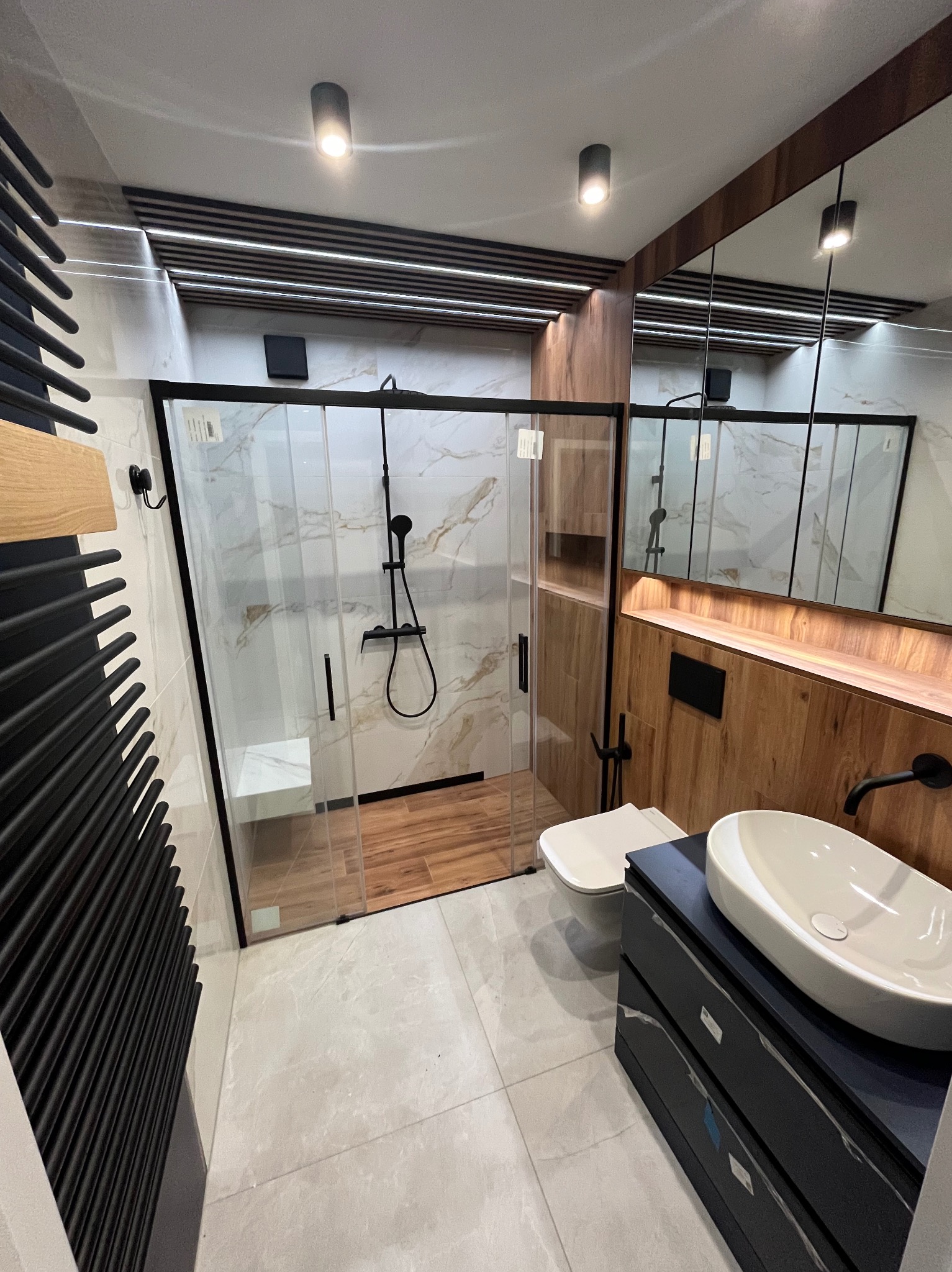 Nowoczesna łazienka z kabiną prysznicową typu walk-in, wykończona płytkami imitującymi marmur, drewnianymi akcentami i czarnymi elementami armatury.