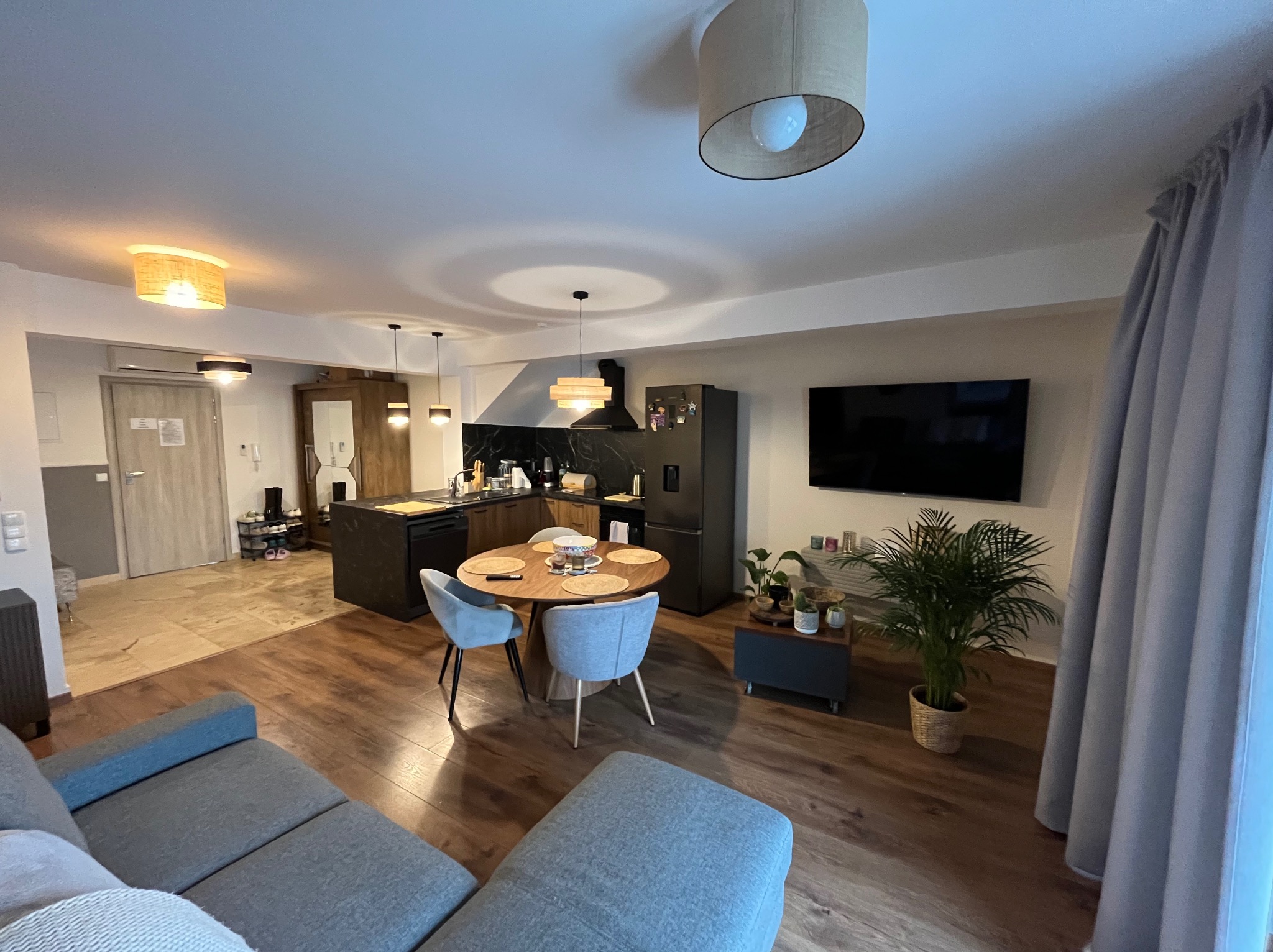 Widok na nowocześnie urządzone wnętrze mieszkania z drewnianą podłogą, szarą sofą, okrągłym stołem z krzesłami oraz aneksem kuchennym z czarnym blatem i oświetleniem punktowym.