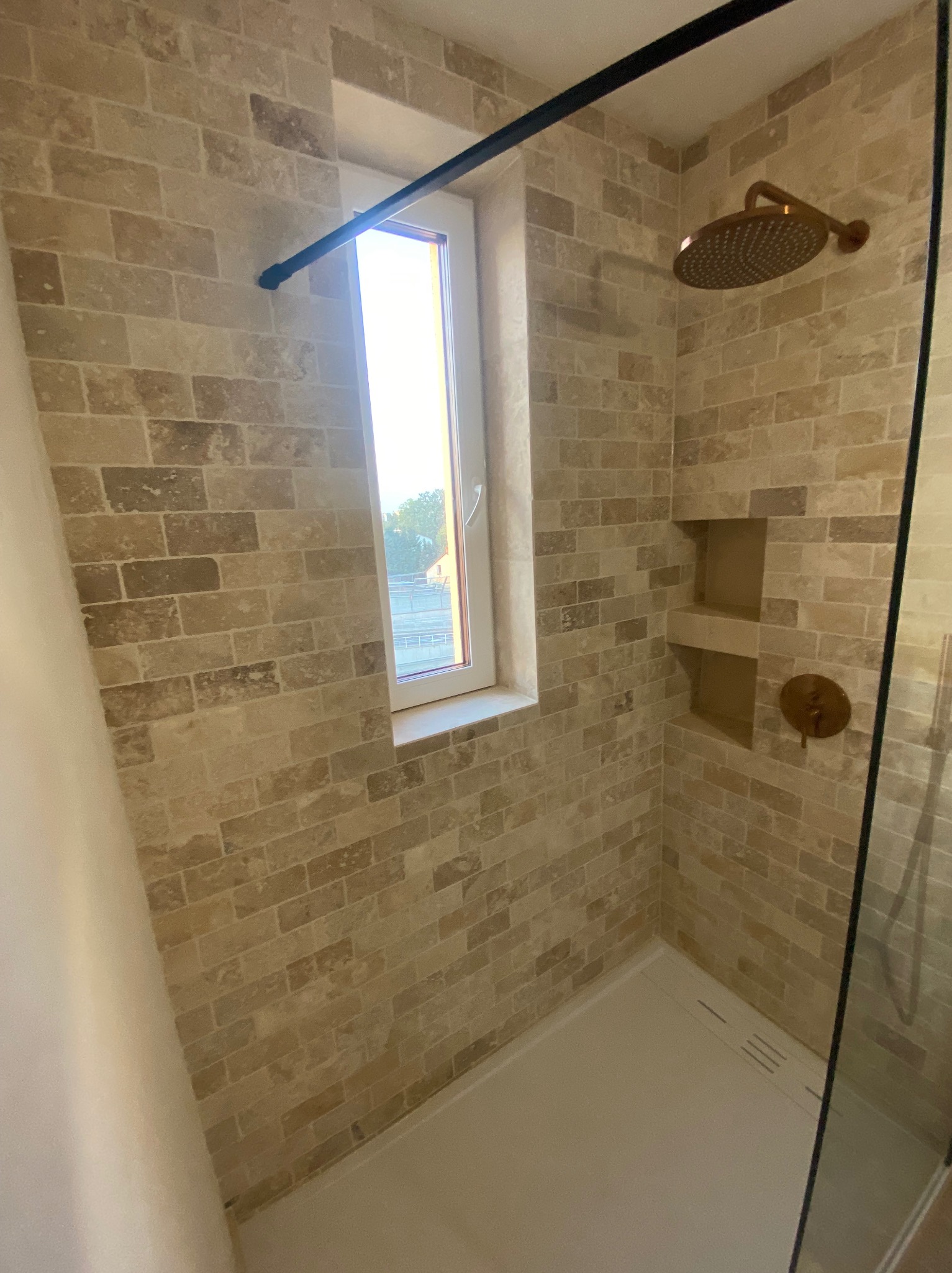 Nowoczesna kabina prysznicowa z beżową mozaiką imitującą cegłę, z wnękami na kosmetyki i deszczownicą w kolorze miedzianym, doświetlona naturalnym światłem z okna.