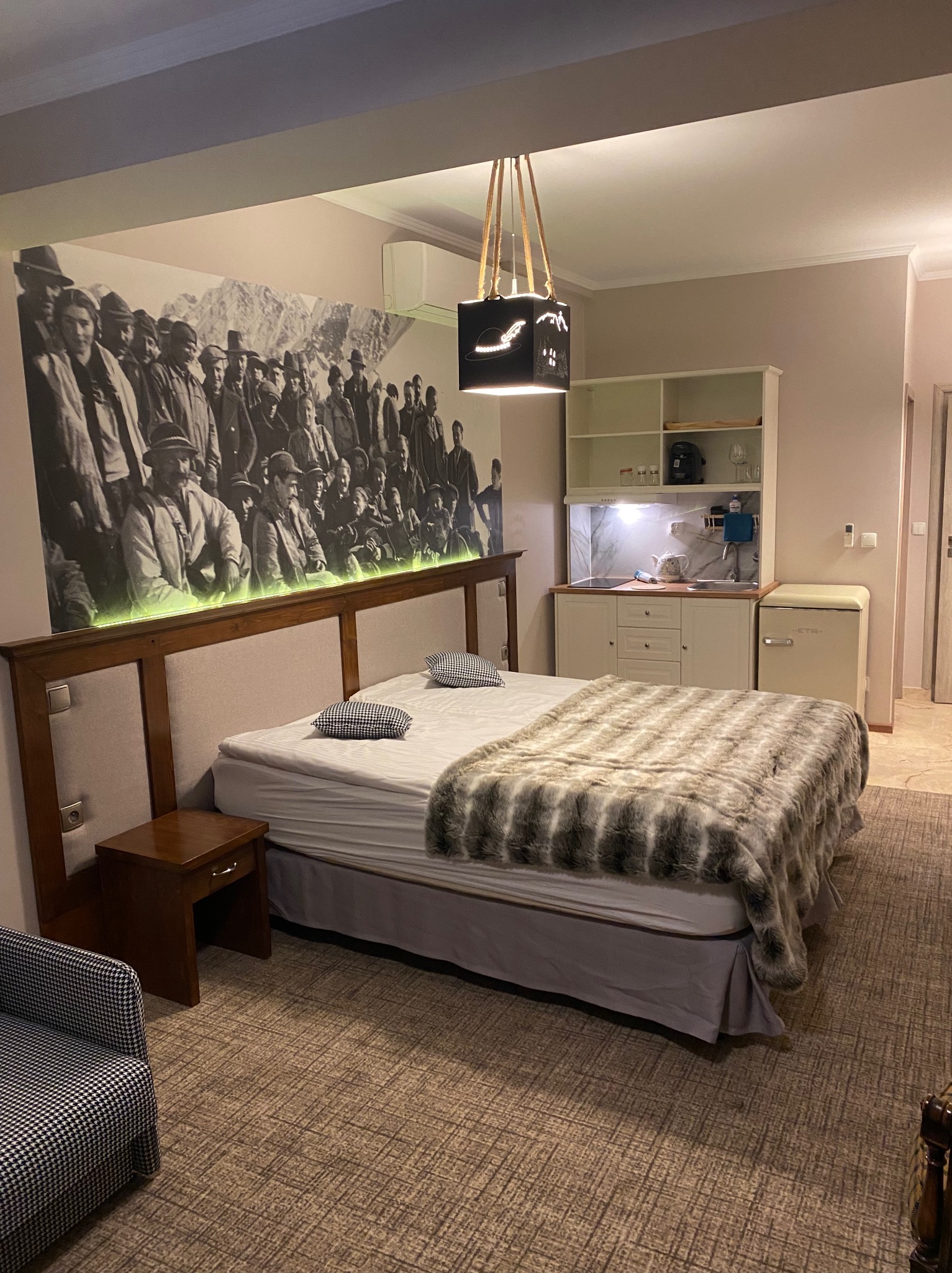 Pokój hotelowy z łóżkiem dwuosobowym, aneksem kuchennym i dużą czarno-białą fotografią na ścianie za wezgłowiem, podświetloną zielonym światłem.