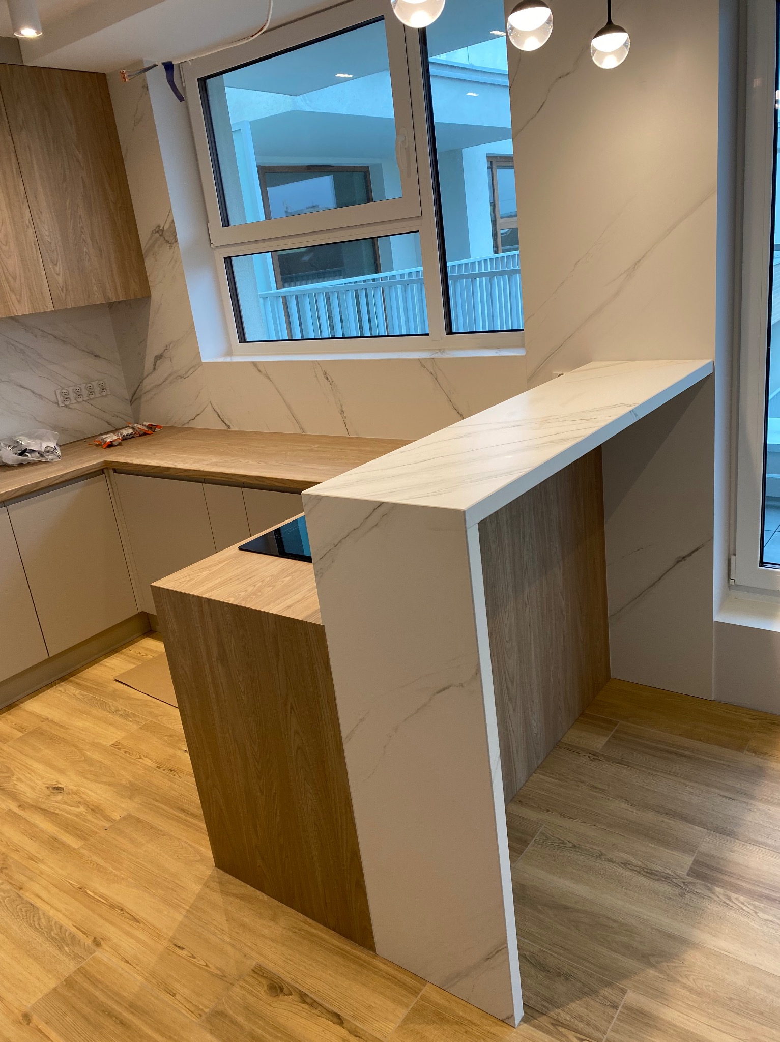 Nowoczesna kuchnia z drewnianymi szafkami, blatem i barkiem wykończonymi w imitacji marmuru, oświetlona wiszącymi lampami, z widokiem na balkon za oknem.