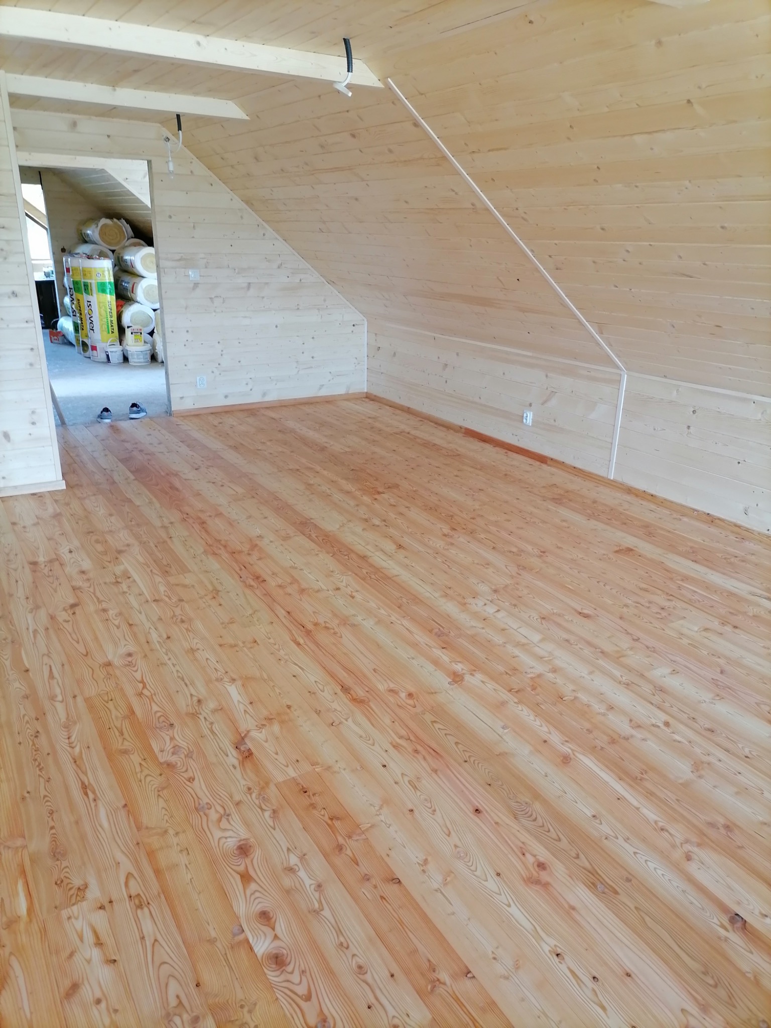 Poddasze wykończone boazerią: drewniana podłoga, ściany i skośny sufit, widoczne materiały izolacyjne w sąsiednim pomieszczeniu.