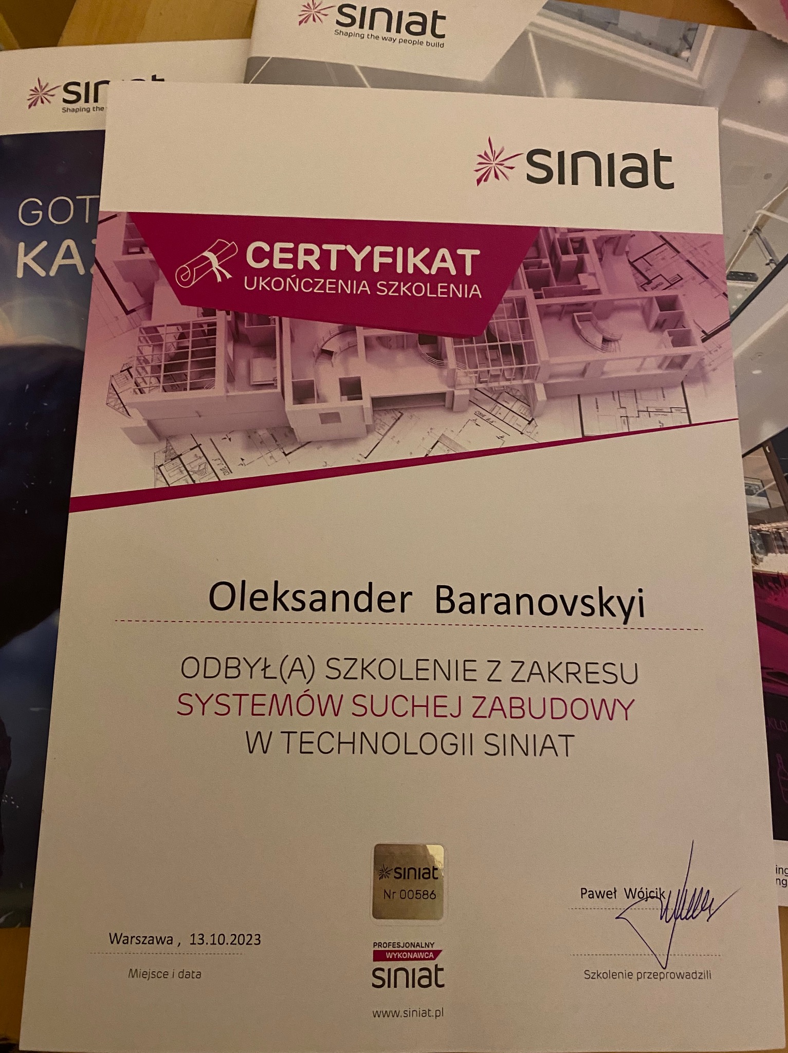 Certyfikat ukończenia szkolenia z zakresu systemów suchej zabudowy w technologii Siniat, wystawiony dla Oleksandra Baranovskyiego, z planem architektonicznym w tle, podpisany przez Pawła Wójcika...