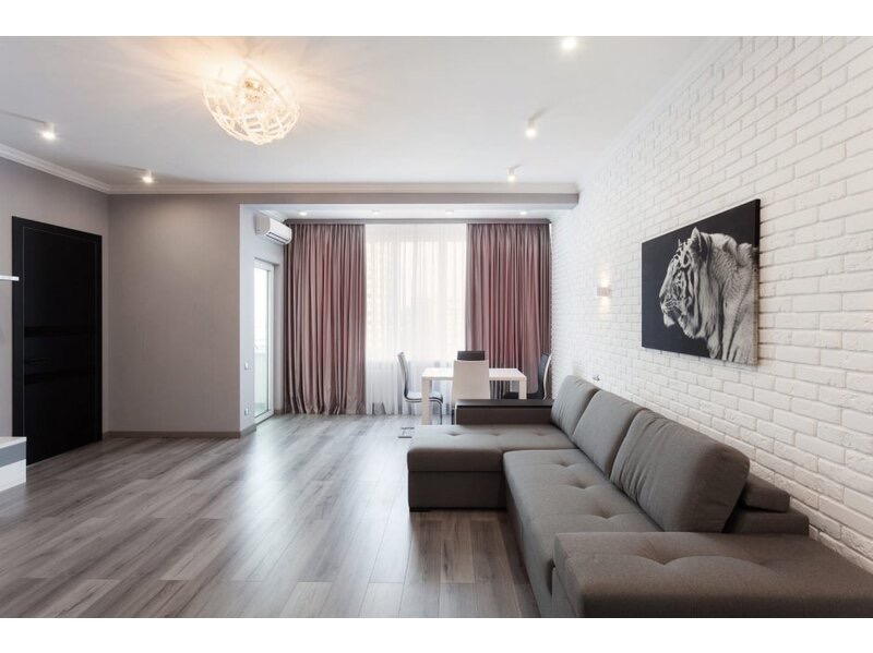 Jasne wnętrze pokoju z szarą, modułową sofą, białą ścianą z cegły, obrazem tygrysa, drewnianą podłogą, okrągłą lampą sufitową i zasłonami w odcieniach różu.