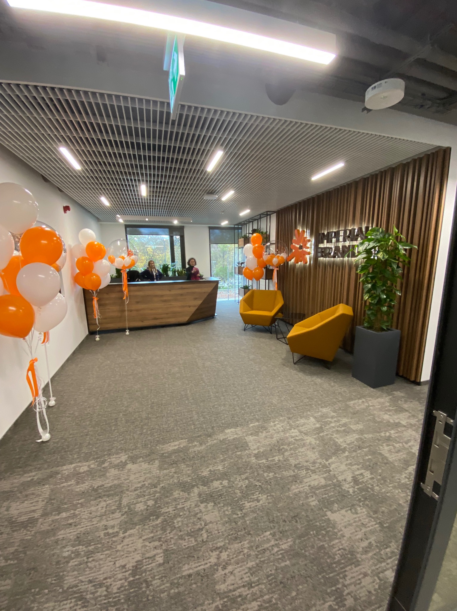 Nowoczesne biuro z recepcją, drewnianą ścianą z logo firmy Imperial Brands, dwoma żółtymi fotelami i dekoracją z białych i pomarańczowych balonów.