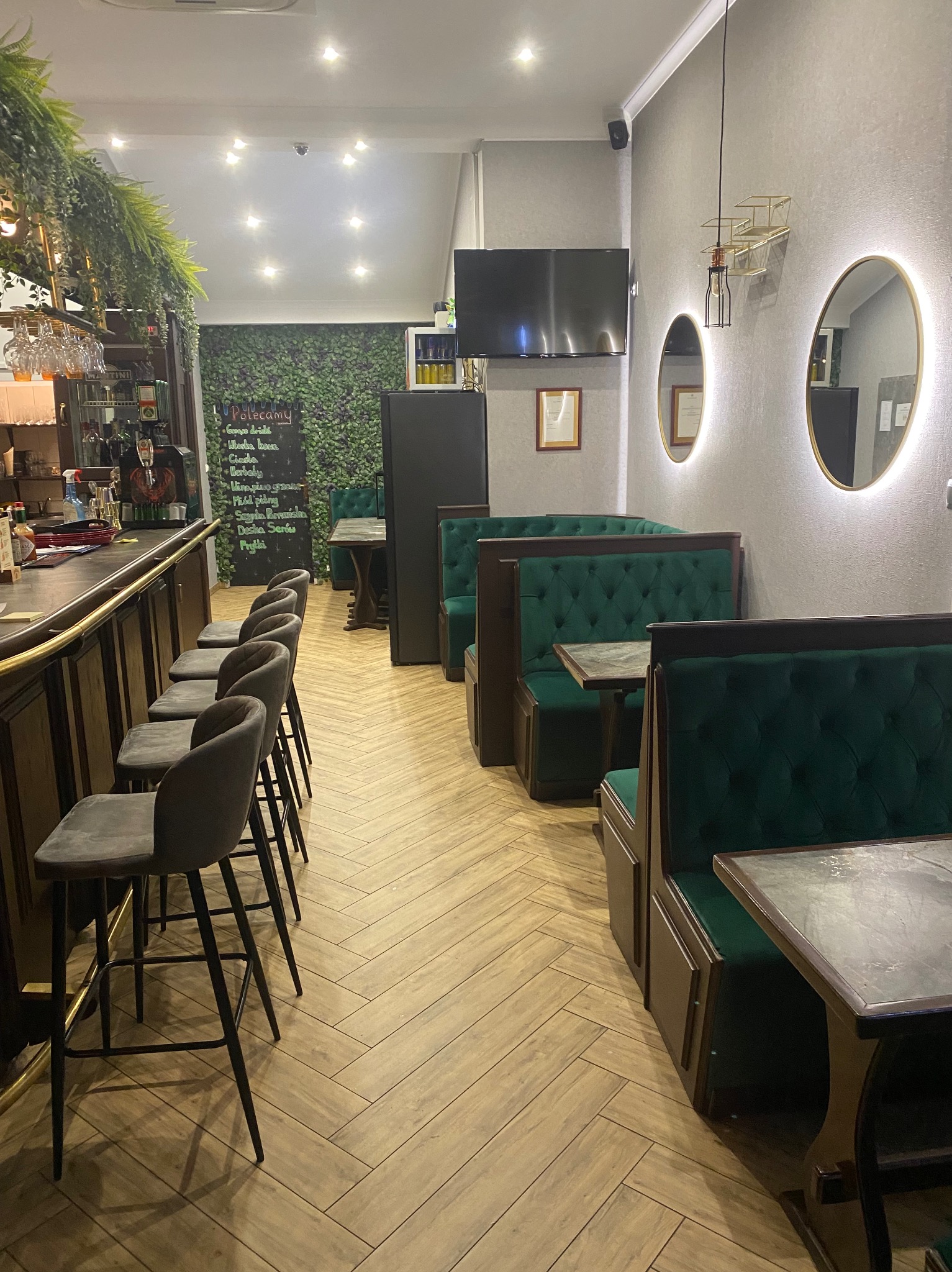 Wnętrze lokalu gastronomicznego z baru z wysokimi krzesłami i zielonymi lożami, parkiet ułożony w jodełkę, dekoracyjna ściana z roślinnością i podświetlane okrągłe lustra.