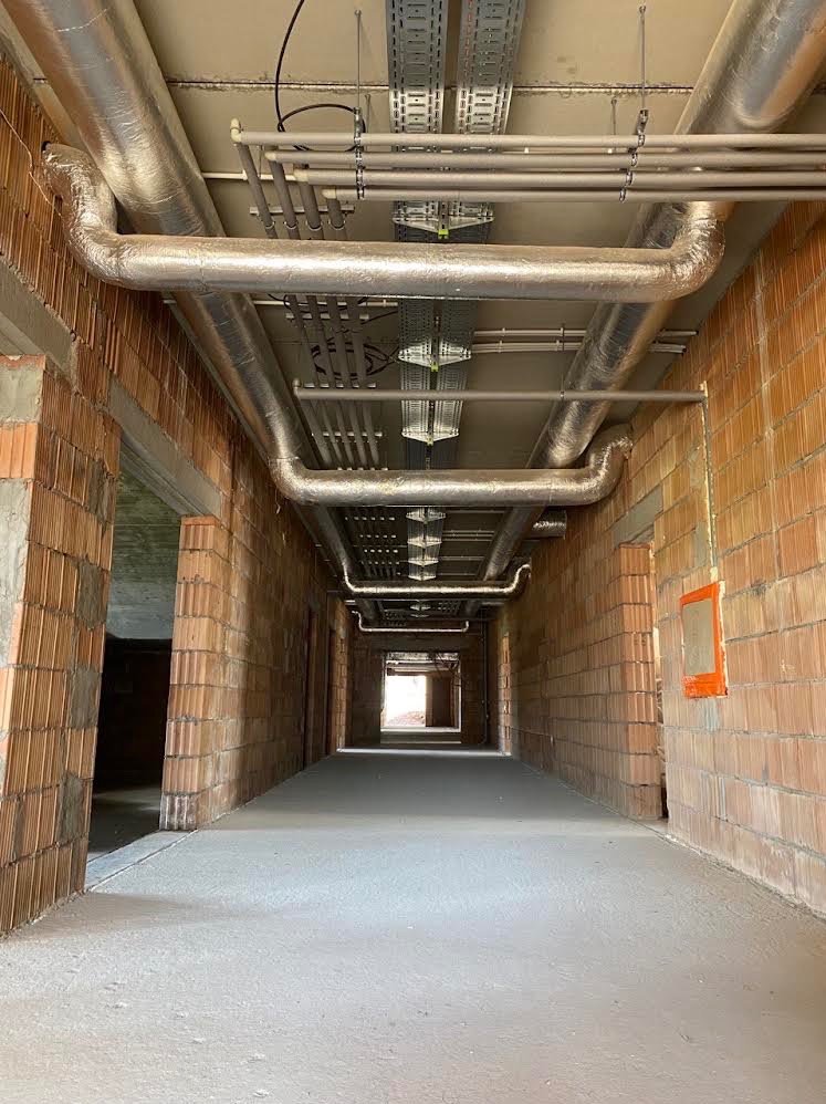 Korytarz w surowym stanie z widocznymi instalacjami wentylacyjnymi i elektrycznymi pod sufitem, ściany z cegły dziurawki, w oddali widoczne światło dzienne.