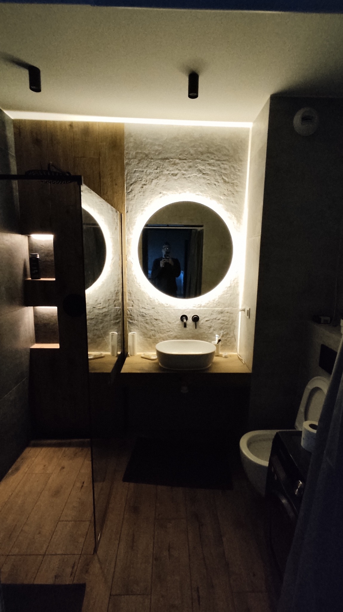 Nowoczesna łazienka z okrągłym lustrem podświetlanym LED, umywalką nablatową i drewnianą podłogą, widoczne elementy wyposażenia i armatury w ciemnych kolorach.