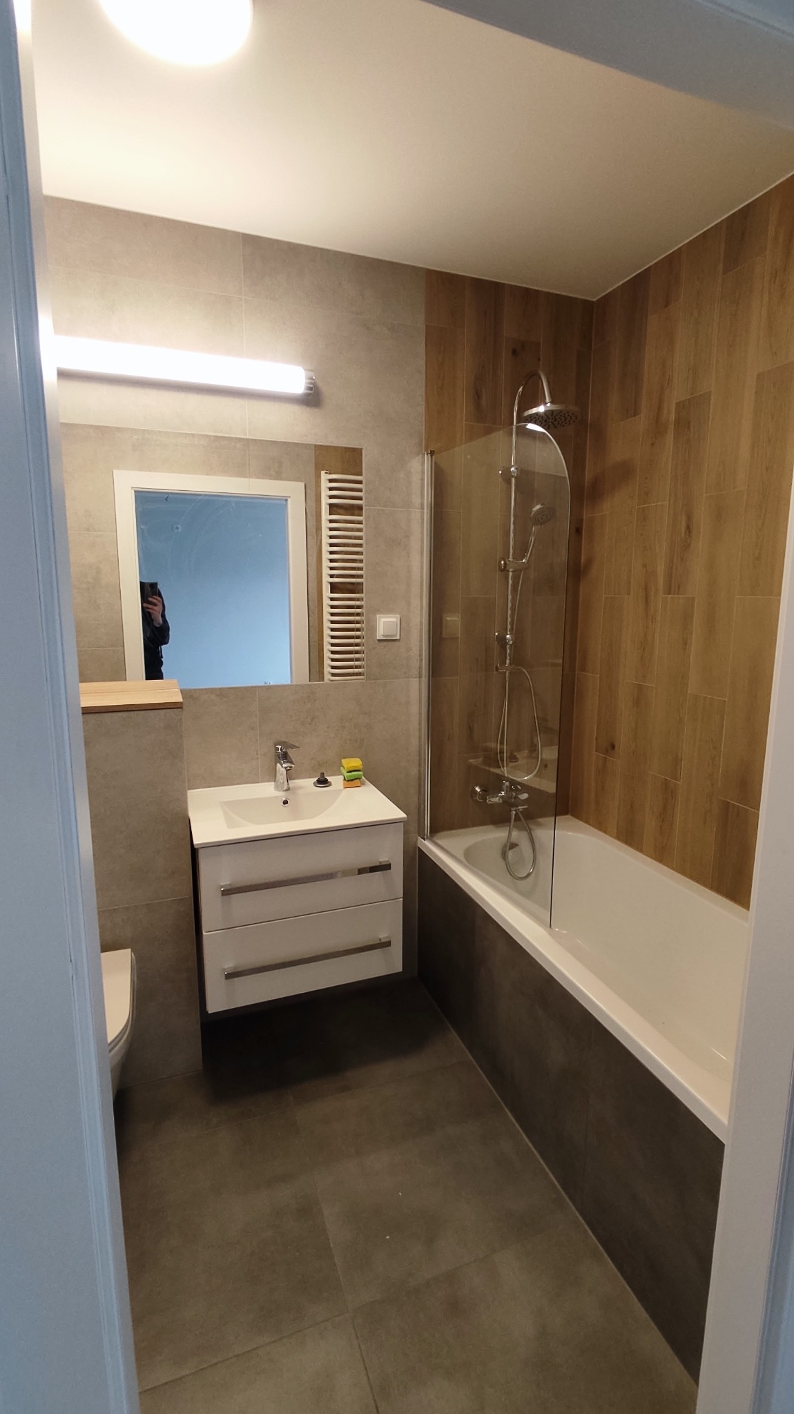 Nowoczesna łazienka po renowacji z drewnopodobnymi panelami na ścianie przy wannie, szklanym parawanem nawannowym, umywalką z szafką w kolorze białym, dużym lustrem z oświetleniem i grzejnikiem...