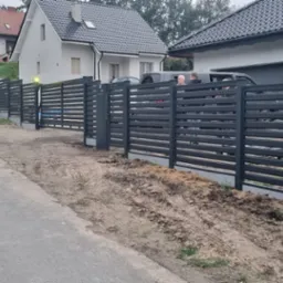Nowoczesne ogrodzenie posesji z poziomych, stalowych profili w kolorze grafitowym, widok z ulicy, w tle domy jednorodzinne.