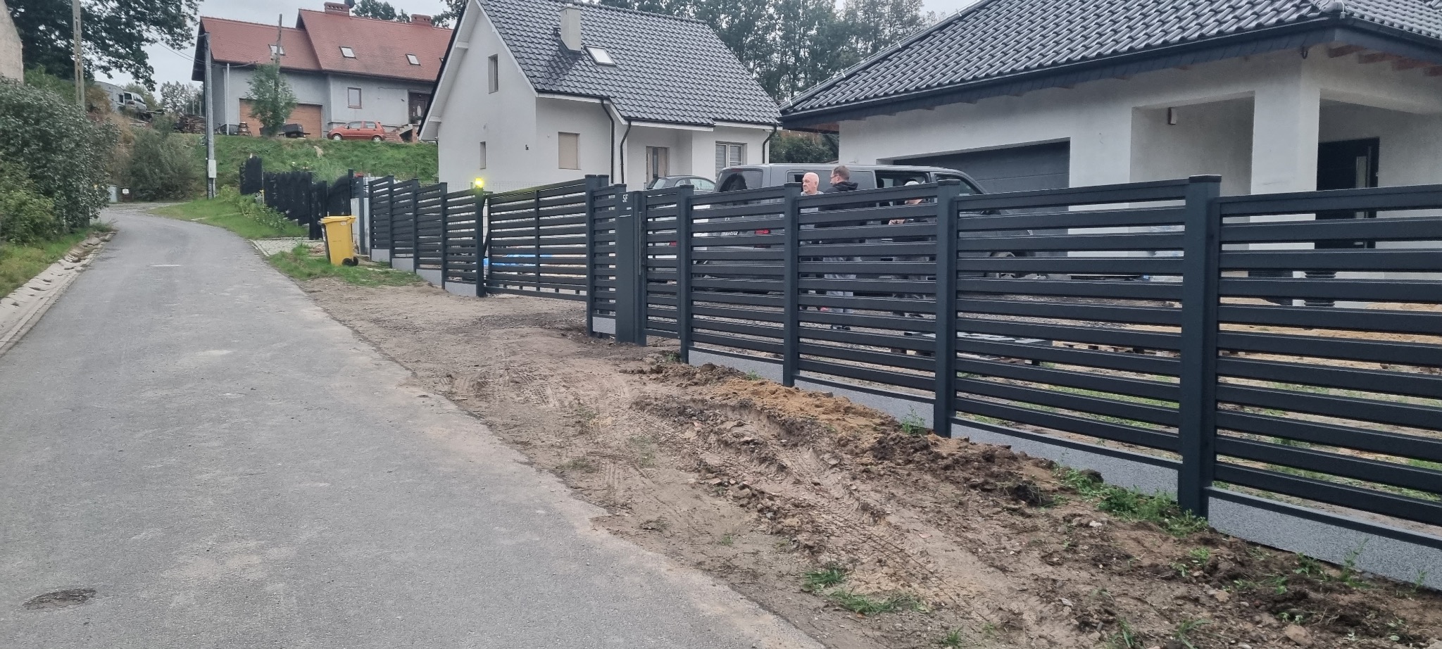 Nowoczesne ogrodzenie posesji z poziomych, stalowych profili w kolorze grafitowym, widok z ulicy, w tle domy jednorodzinne.