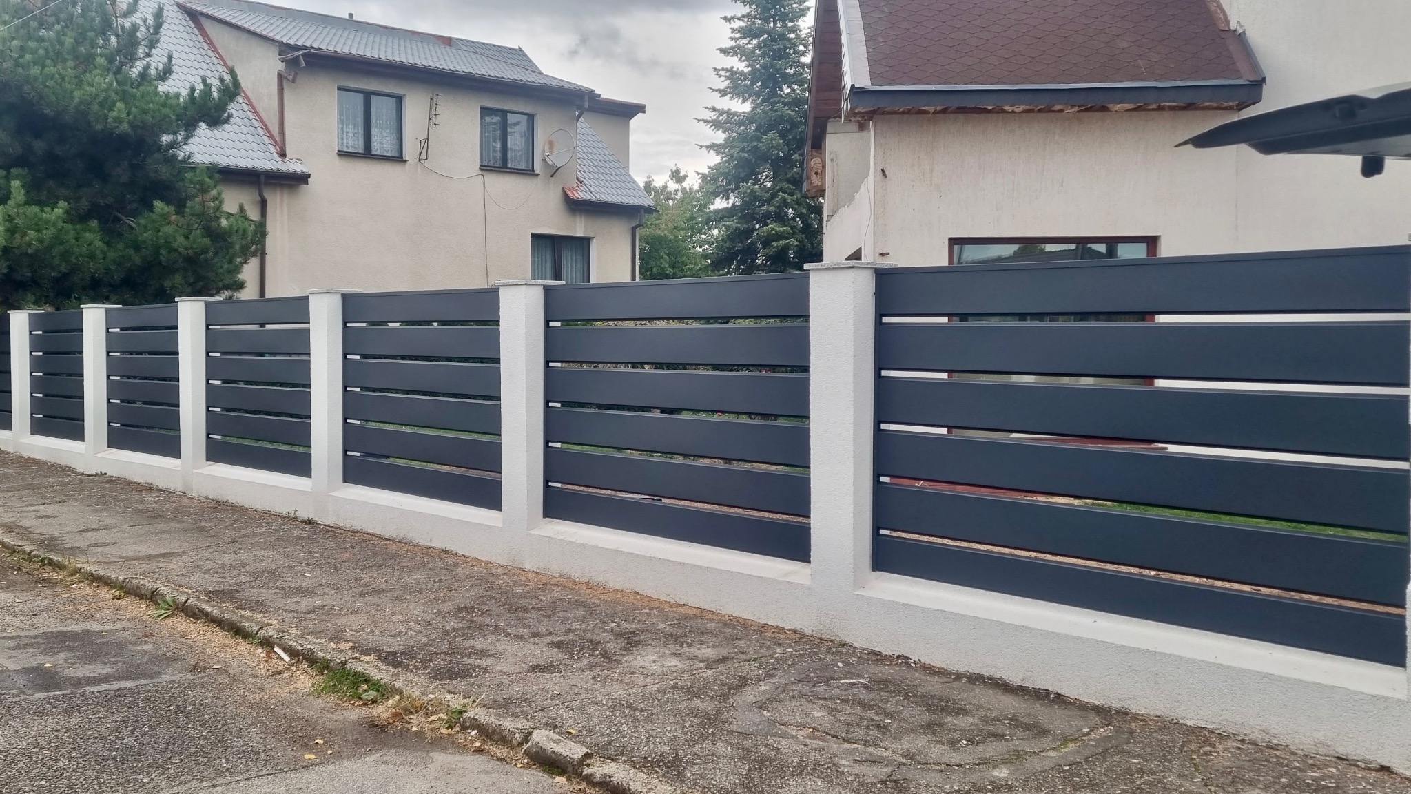 Nowoczesne ogrodzenie poziome z szarych paneli zamontowanych na białych, murowanych słupkach i podmurówce. Widok z poziomu chodnika na tle dwóch domów.