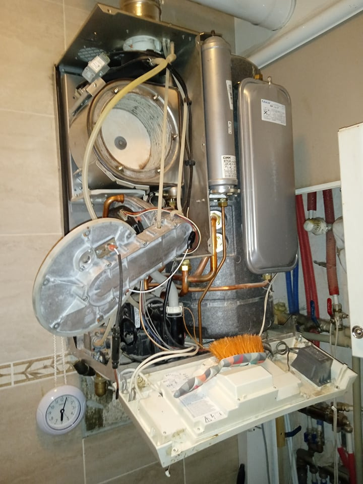 Otwarty piec gazowy z widocznymi elementami wewnętrznymi, obok leży szczotka, a na ścianie wisi zegar.