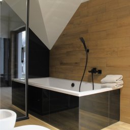 FARMA Styl Elżbieta Wieczorek - Nowoczesna łazienka z wanną obudowaną czarnymi płytkami imitującymi marmur, drewnopodobne płytki na ścianie i podłodze, czarna armatura i szklana kabina.