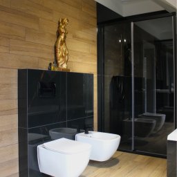 FARMA Styl Elżbieta Wieczorek - Nowoczesna łazienka z drewnianymi i czarnymi płytkami, białą armaturą (toaleta i bidet) oraz złotą figurką. Kabina prysznicowa z czarnymi ramami.