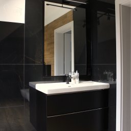 FARMA Styl Elżbieta Wieczorek - Nowoczesna łazienka z czarnymi płytkami imitującymi marmur, białą umywalką nablatową i dużym lustrem z oświetleniem LED. Widoczne drewniane akcenty w odbiciu lustra.