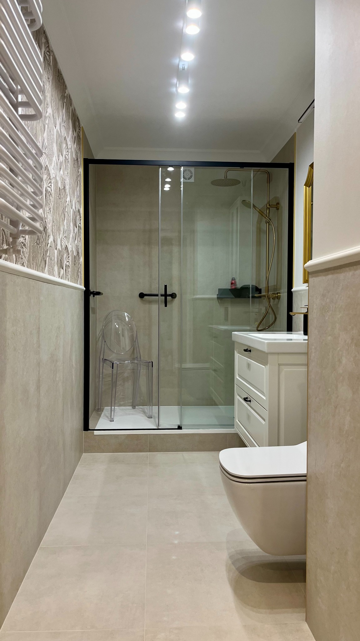 Jasna łazienka z kabiną prysznicową w czarnej ramie, umywalką z szafką w stylu retro i wiszącą toaletą. Na ścianie wzorzysta tapeta i grzejnik drabinkowy.