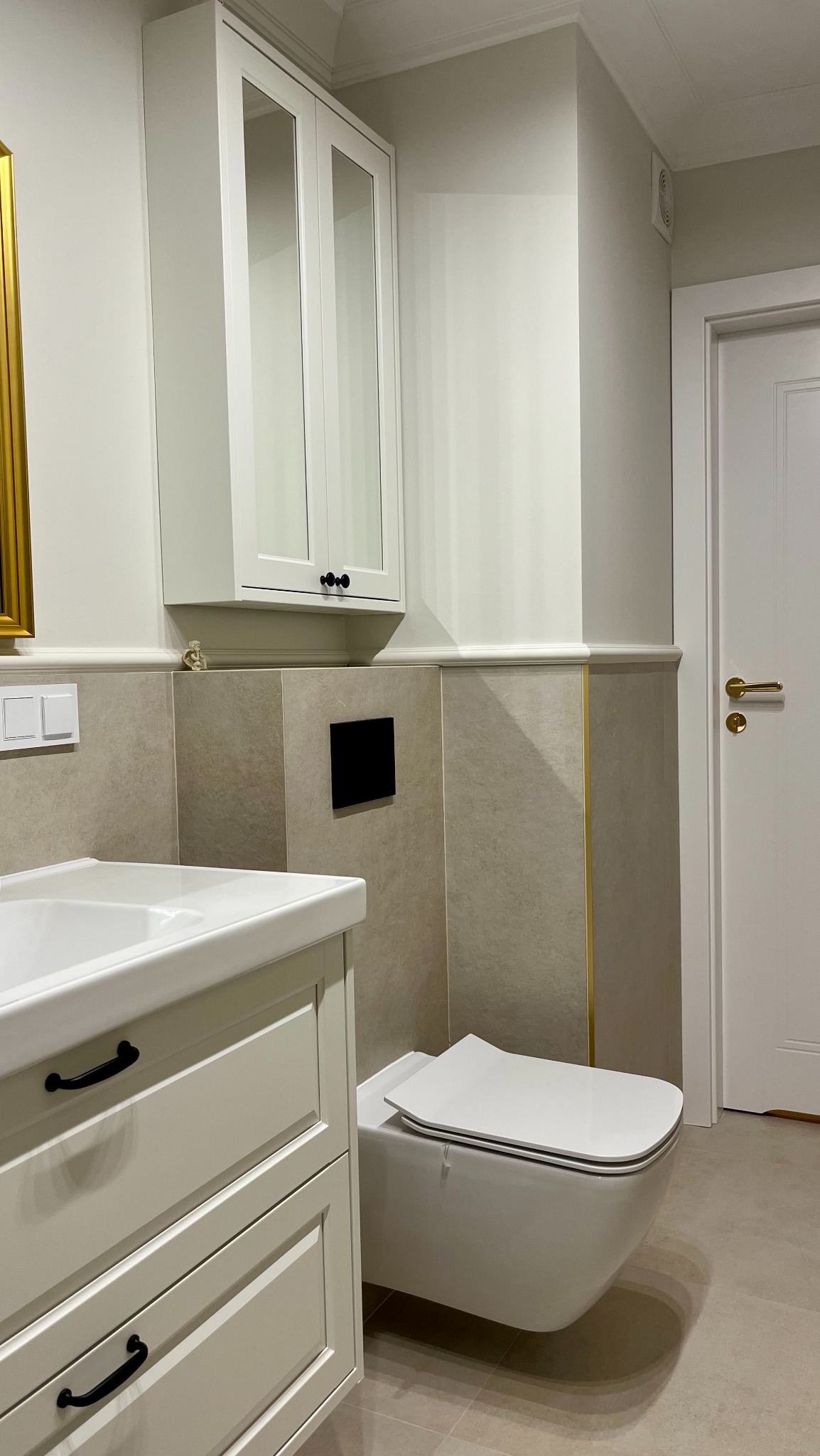 Jasna łazienka w stylu klasycznym: biała szafka z umywalką, podwieszana toaleta, płytki w odcieniach beżu z delikatnym złotym akcentem. Minimalistyczny design.