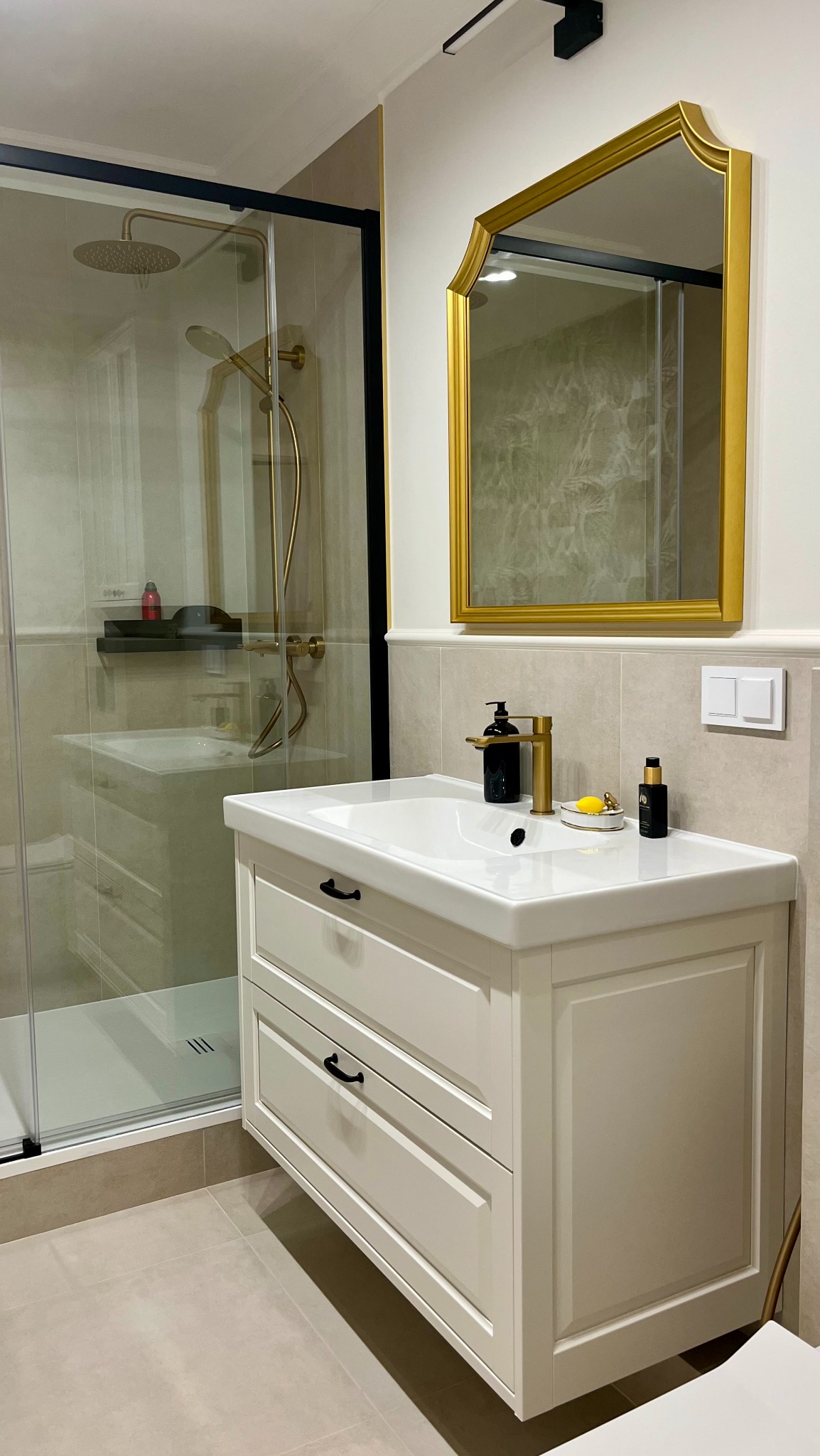 Elegancka łazienka: biała szafka z umywalką, złota armatura i rama lustra, kabina prysznicowa z czarnymi ramami. Minimalistyczny design, beżowe płytki.