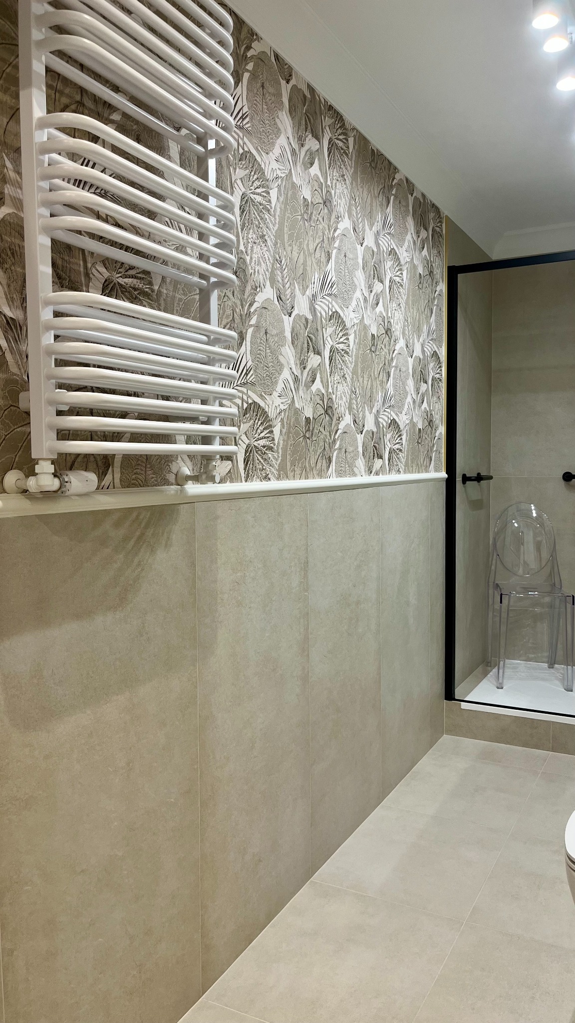 Beżowa łazienka z grzejnikiem drabinkowym, wzorzystą tapetą w liście i przezroczystym krzesłem w kabinie prysznicowej. Minimalistyczny design.