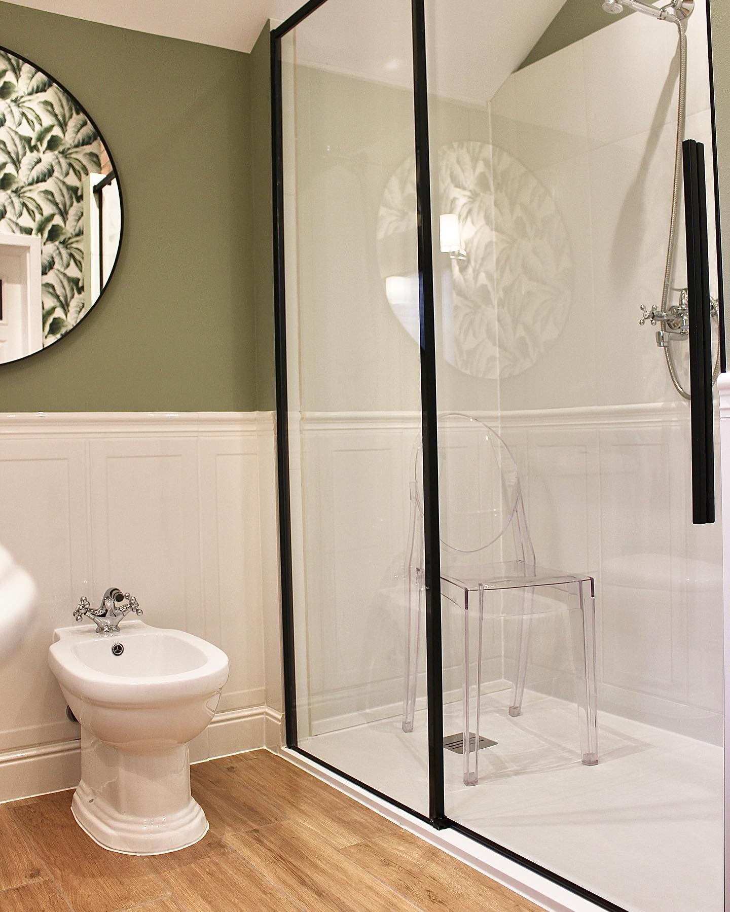 Nowoczesna łazienka z bidetem, kabiną prysznicową z czarnymi ramami i przezroczystym krzesłem. Lustro w okrągłej ramie z motywem roślinnym.