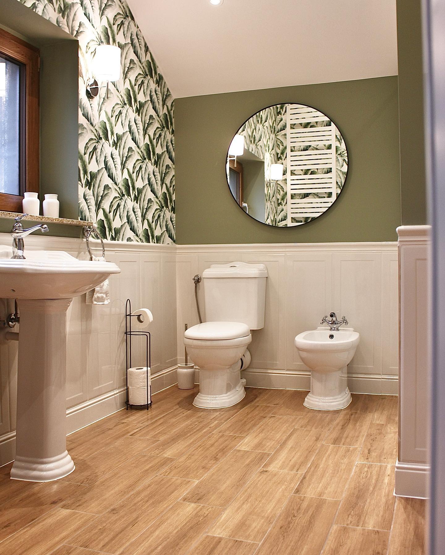 Jasna łazienka w stylu retro z drewnianą podłogą, umywalką na postumencie, toaletą i bidetem. Ściany zdobią białe panele i tapeta w liście. Okrągłe lustro.