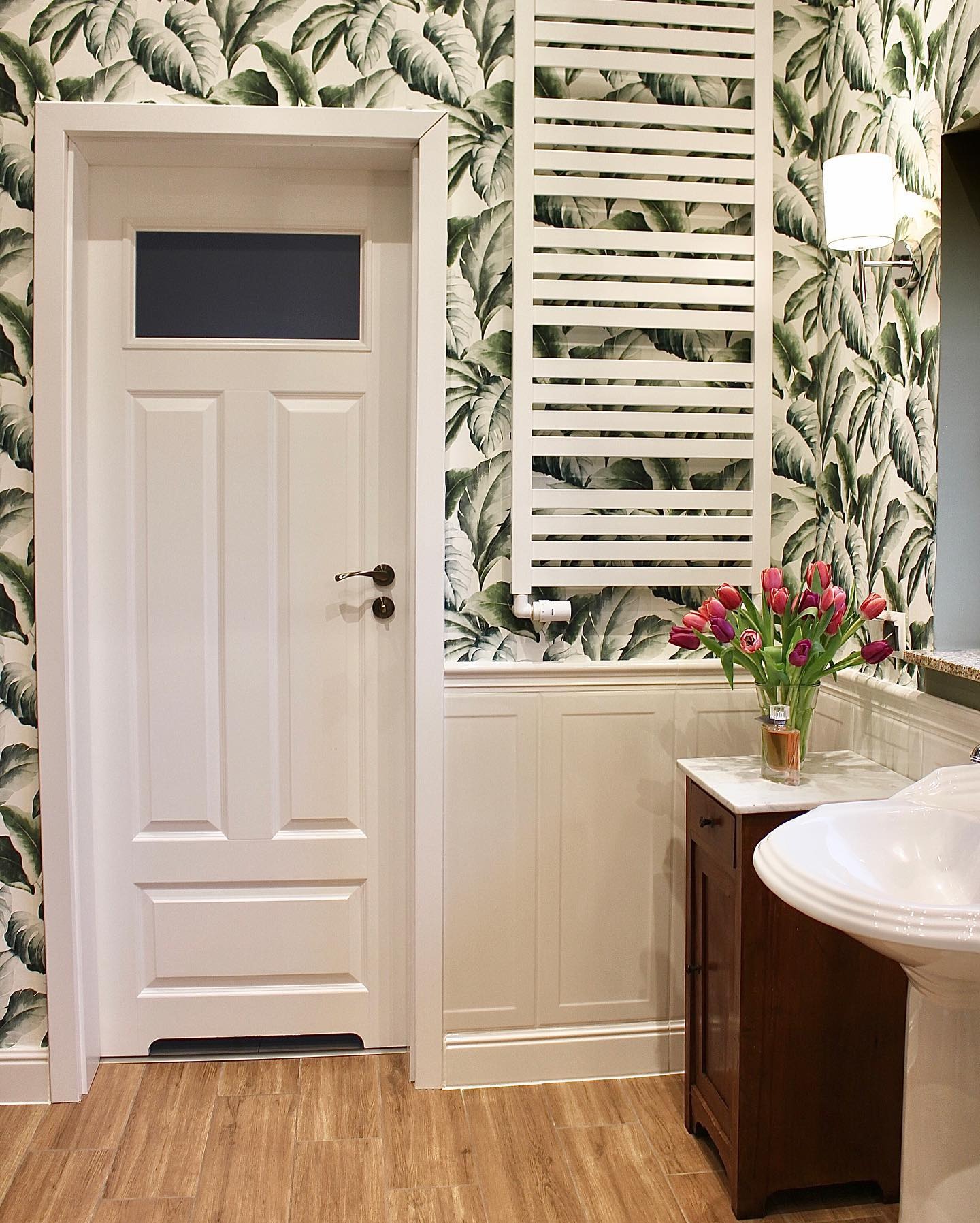 Biała łazienka z motywem tropikalnych liści na tapecie, grzejnikiem drabinkowym i umywalką na postumencie. Na szafce stoi wazon z tulipanami i flakon perfum.