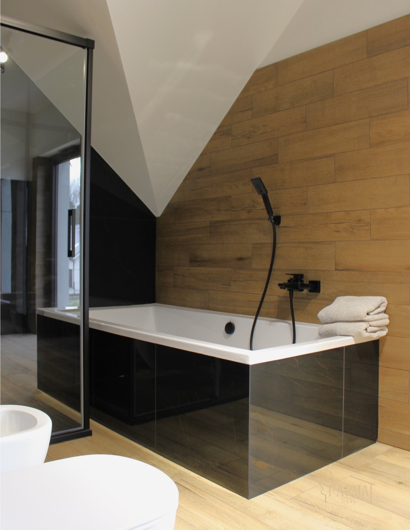Nowoczesna łazienka z wanną obudowaną czarnymi płytkami imitującymi marmur, drewnopodobne płytki na ścianie i podłodze, czarna armatura i szklana kabina.