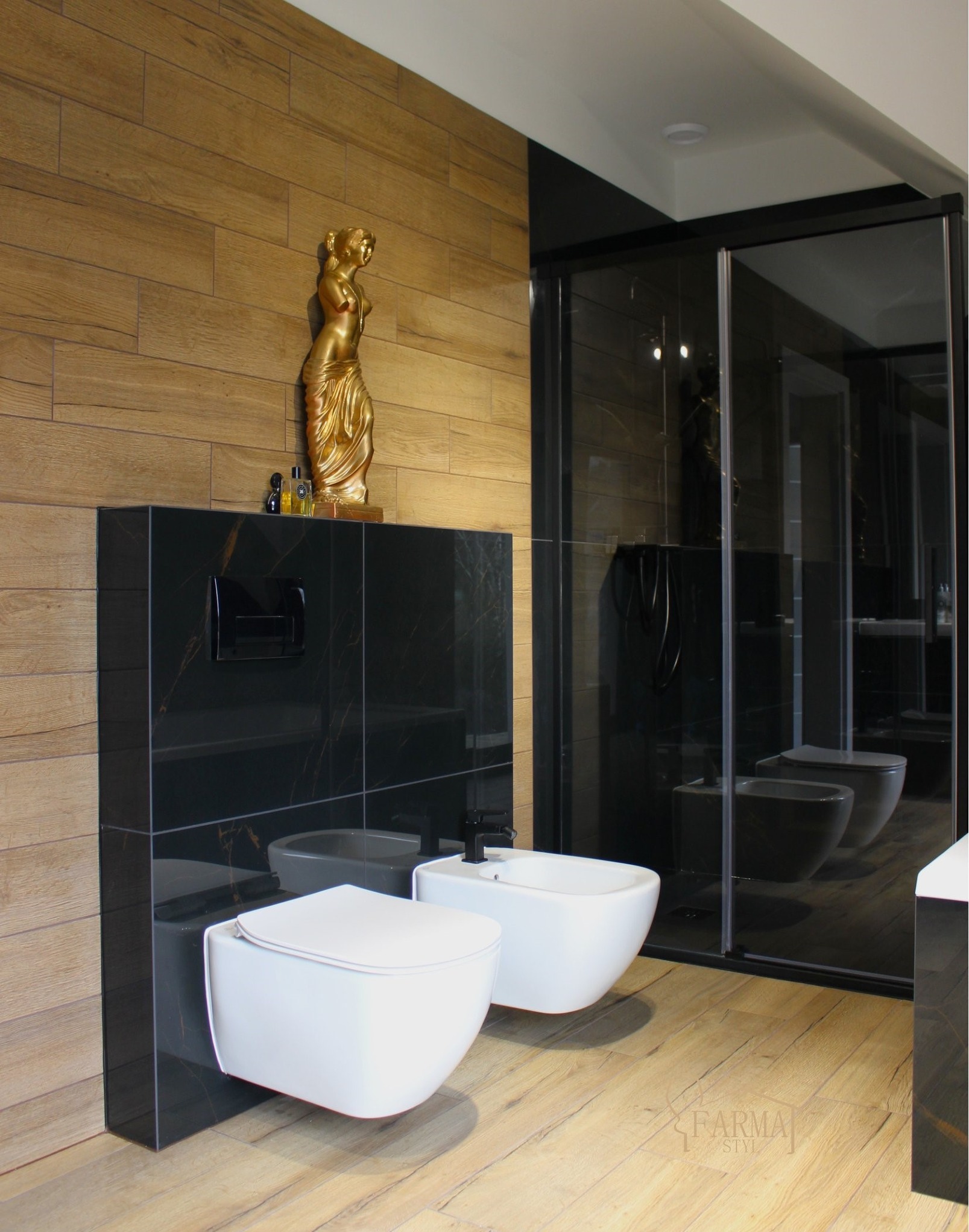 Nowoczesna łazienka z drewnianymi i czarnymi płytkami, białą armaturą (toaleta i bidet) oraz złotą figurką. Kabina prysznicowa z czarnymi ramami.