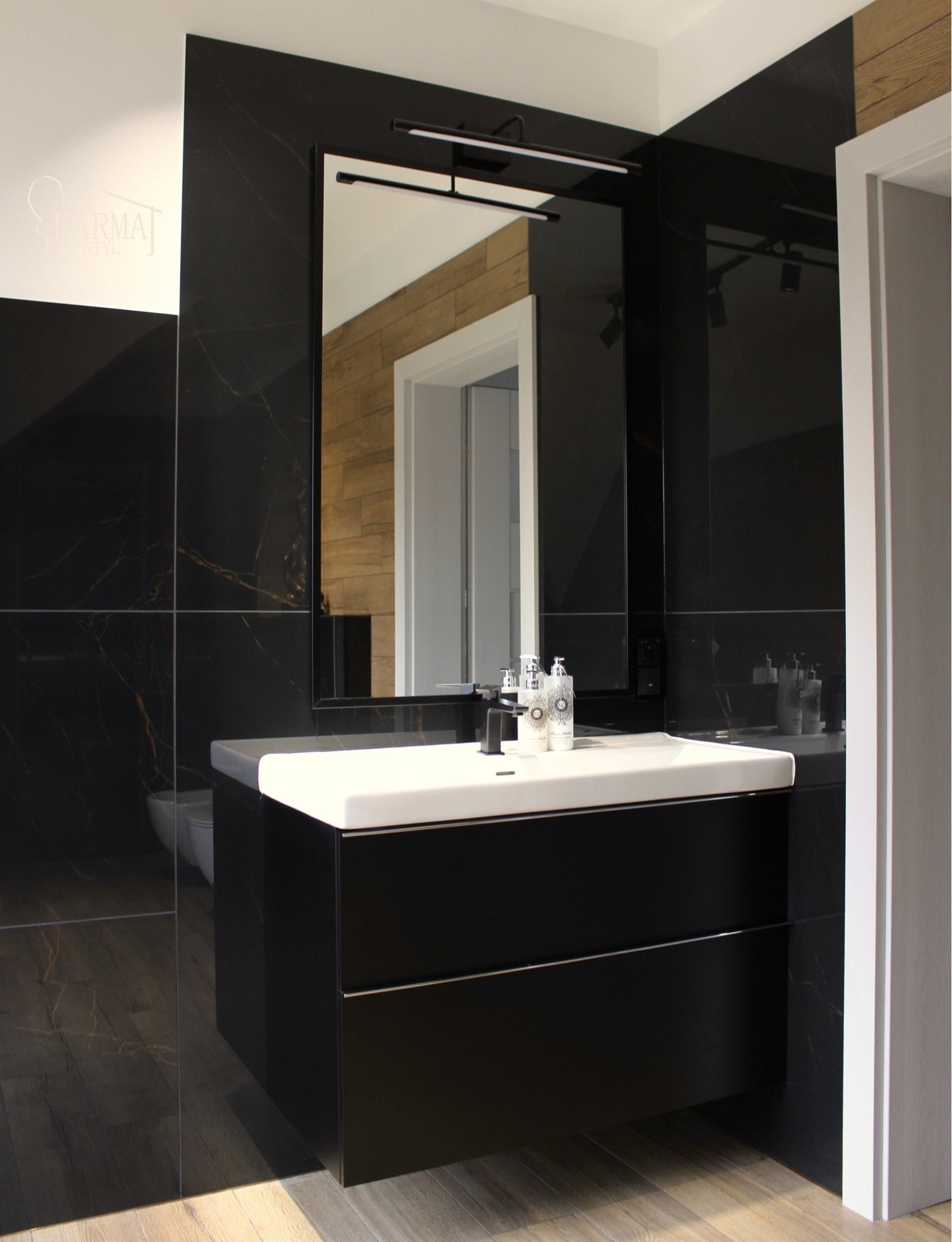 Nowoczesna łazienka z czarnymi płytkami imitującymi marmur, białą umywalką nablatową i dużym lustrem z oświetleniem LED. Widoczne drewniane akcenty w odbiciu lustra.