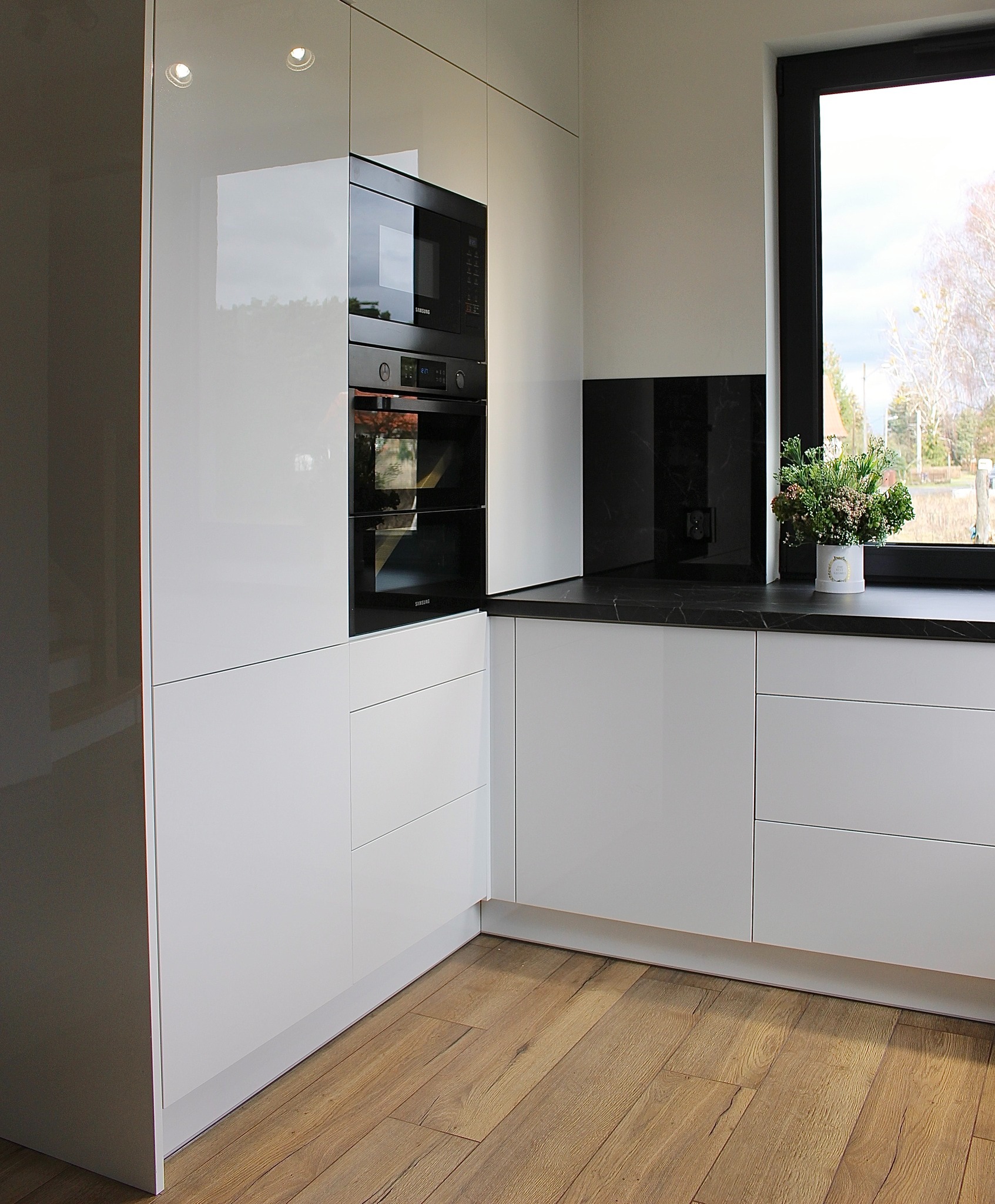 Biała, nowoczesna kuchnia z połyskiem, czarny blat i okno z widokiem na zewnątrz. Minimalistyczny design, drewniana podłoga, kwiaty w doniczce na blacie.